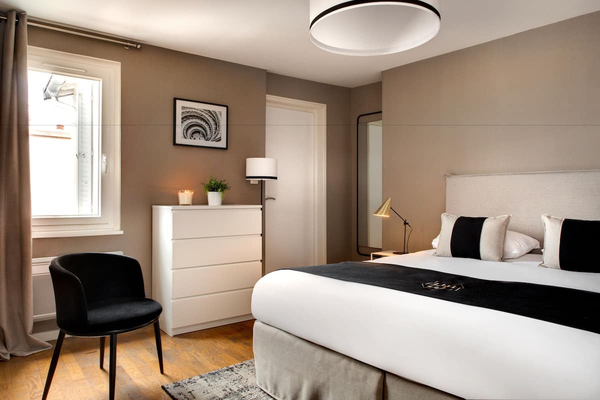 A modern bedroom with a bed, a chair, a dresser, a bedside table with a lamp, a window with curtains, and a wall-mounted picture.