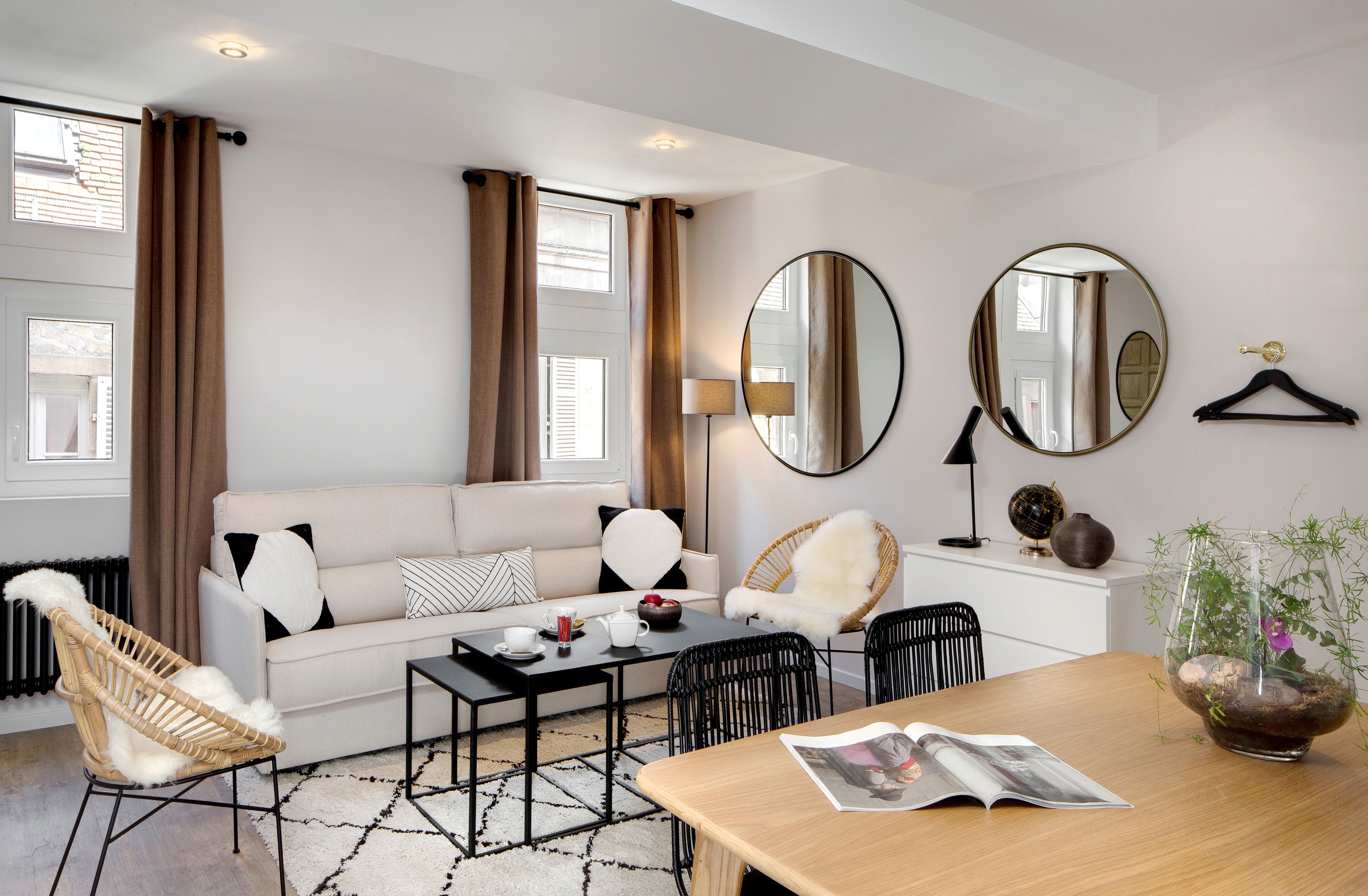 A modern hotel room with a sofa, chairs, a dining table, a lamp, a mirror, and a hanging coat rack.