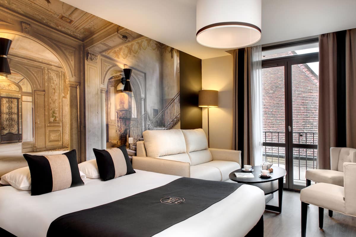 A hotel room with a large bed, two armchairs, a small table, and a view of a building through a large window.
