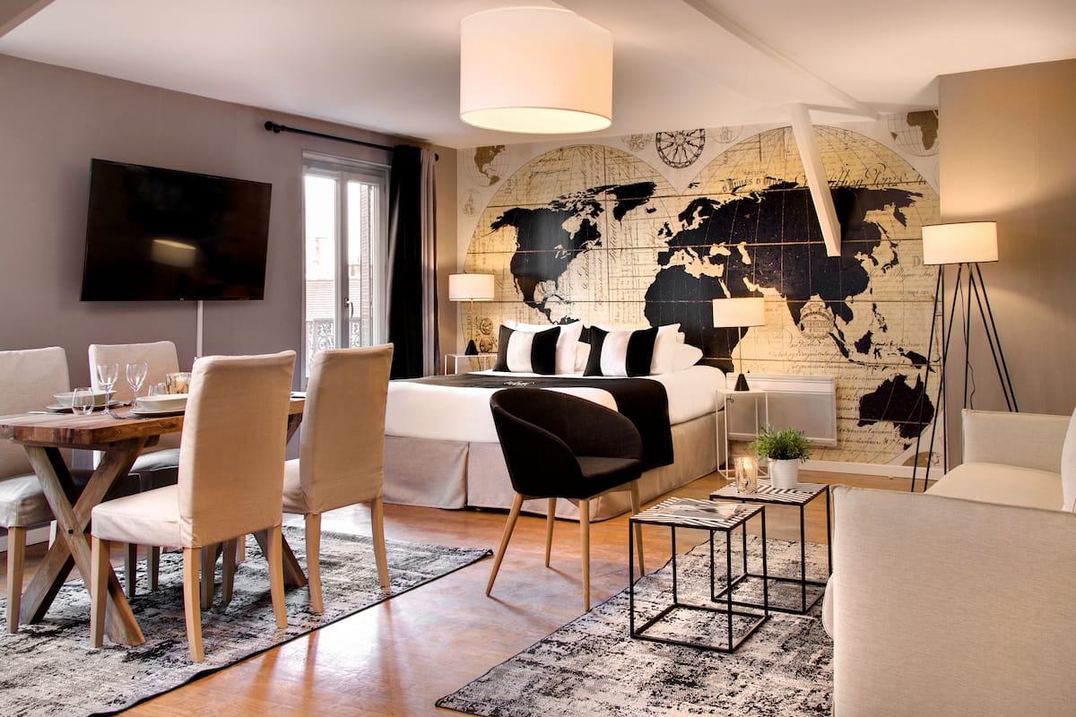 A modern hotel room with a large bed, a dining table with chairs, a TV, and a world map on the wall.