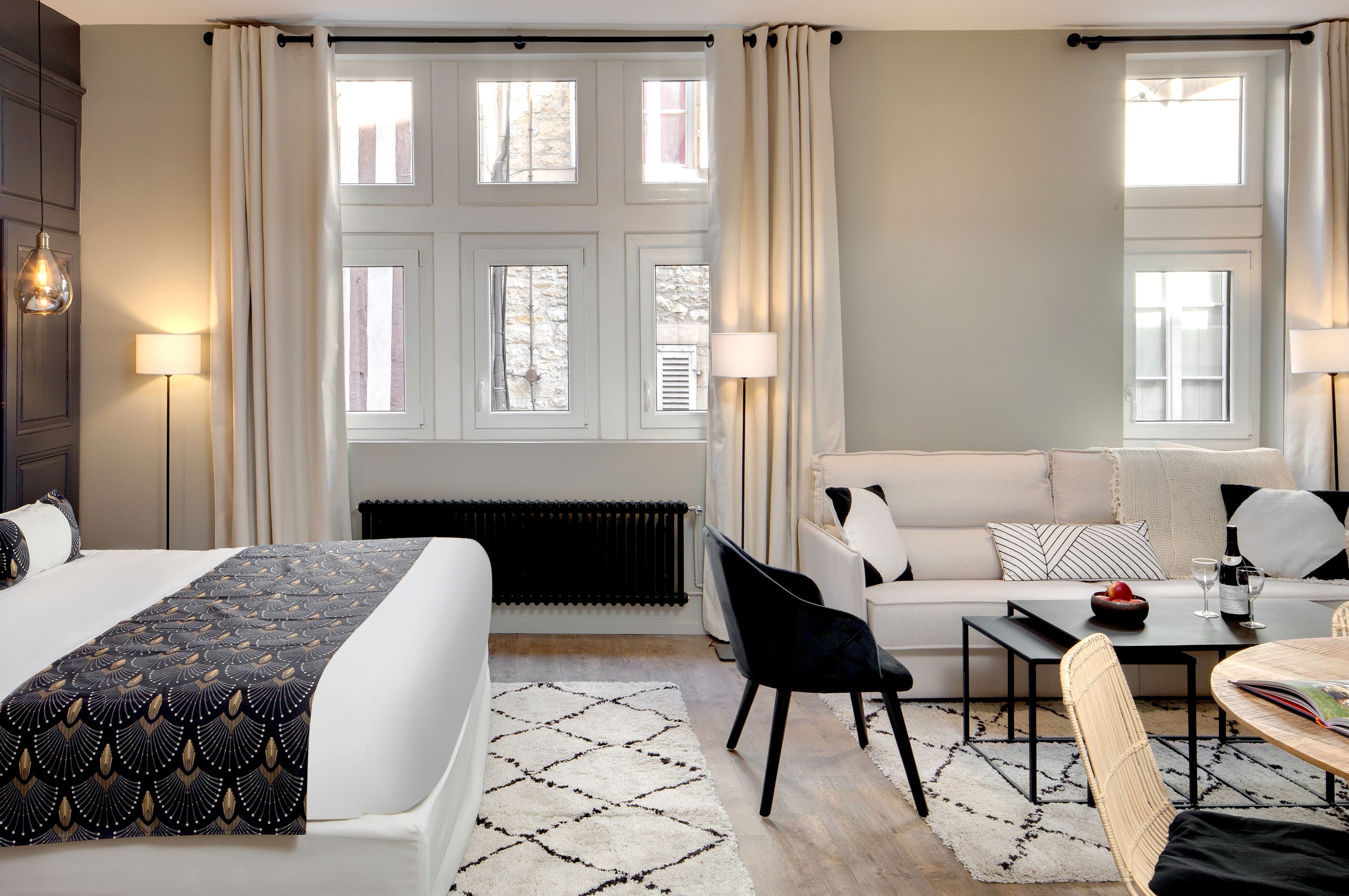 Quarto de hotel com cama, sofá, mesa de jantar e uma cadeira. Há duas janelas com cortinas, um radiador e um abajur no lado esquerdo.