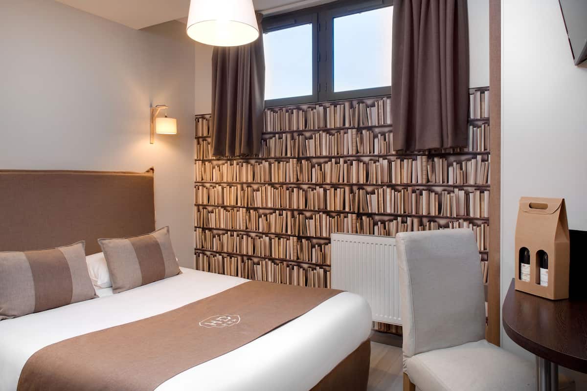 A hotel room with a large bed, a bookshelf wall, a chair, and a table with a bottle of wine.