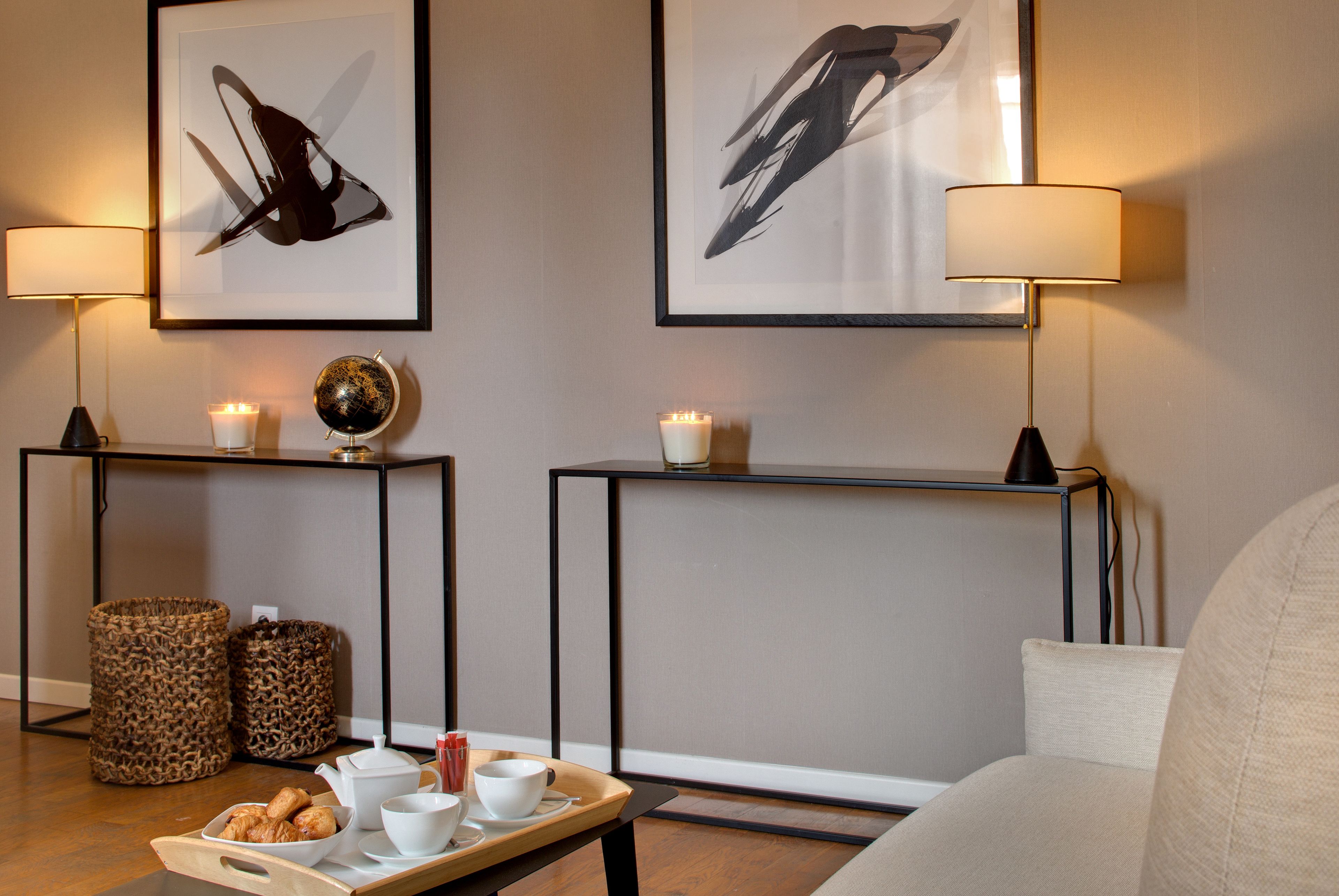 A cozy living area with a sofa, a coffee table with a tray of pastries, two lamps, and two framed abstract paintings on the wall.