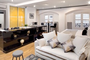 A modern living room with a black kitchen island, white sofa with patterned cushions, and a view of the city through large windows. - Hôtel des Ducs (Dijon)