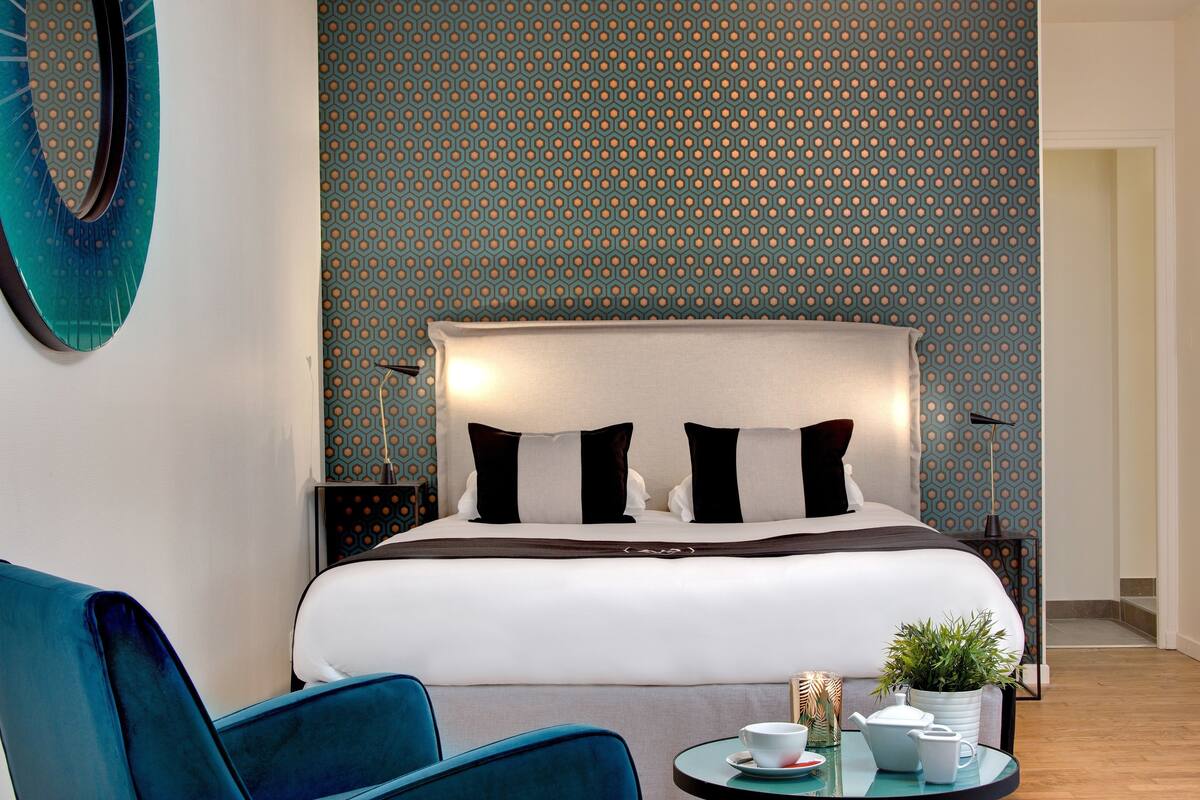 A hotel room with a large bed, two bedside tables, a blue armchair, a small round table with a plant and a teapot, and a patterned wall.