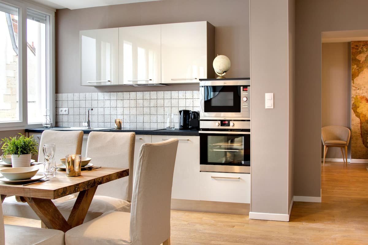A modern kitchen with white cabinets, a microwave, and an oven. There is a dining table with chairs and a potted plant.