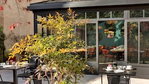 An outdoor dining area with tables and chairs, a large potted plant, and a glass-enclosed dining space with chairs and tables visible through the glass. - Hôtel des Ducs (Dijon)