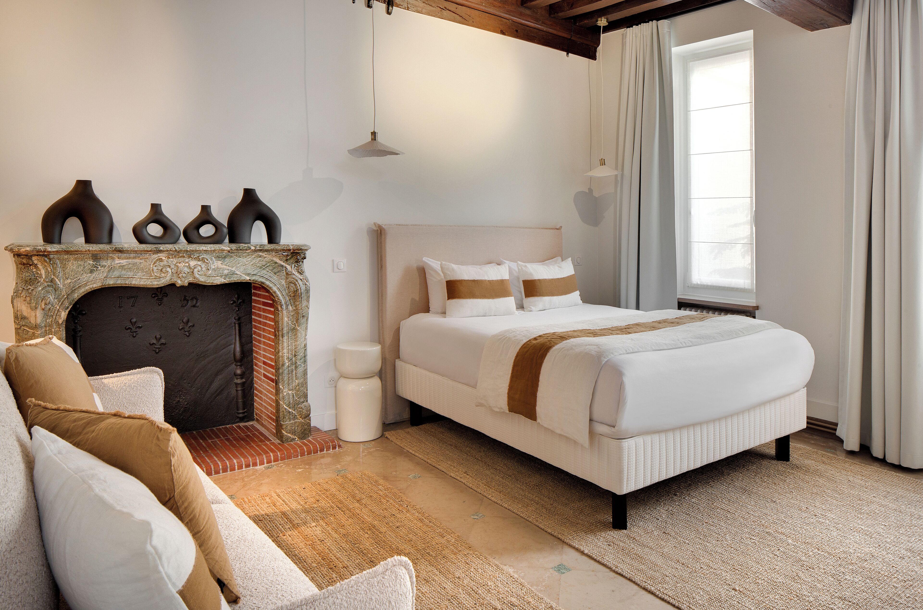 A bedroom with a fireplace, a bed with white bedding and brown accents, a sofa with cushions, and a window with curtains.