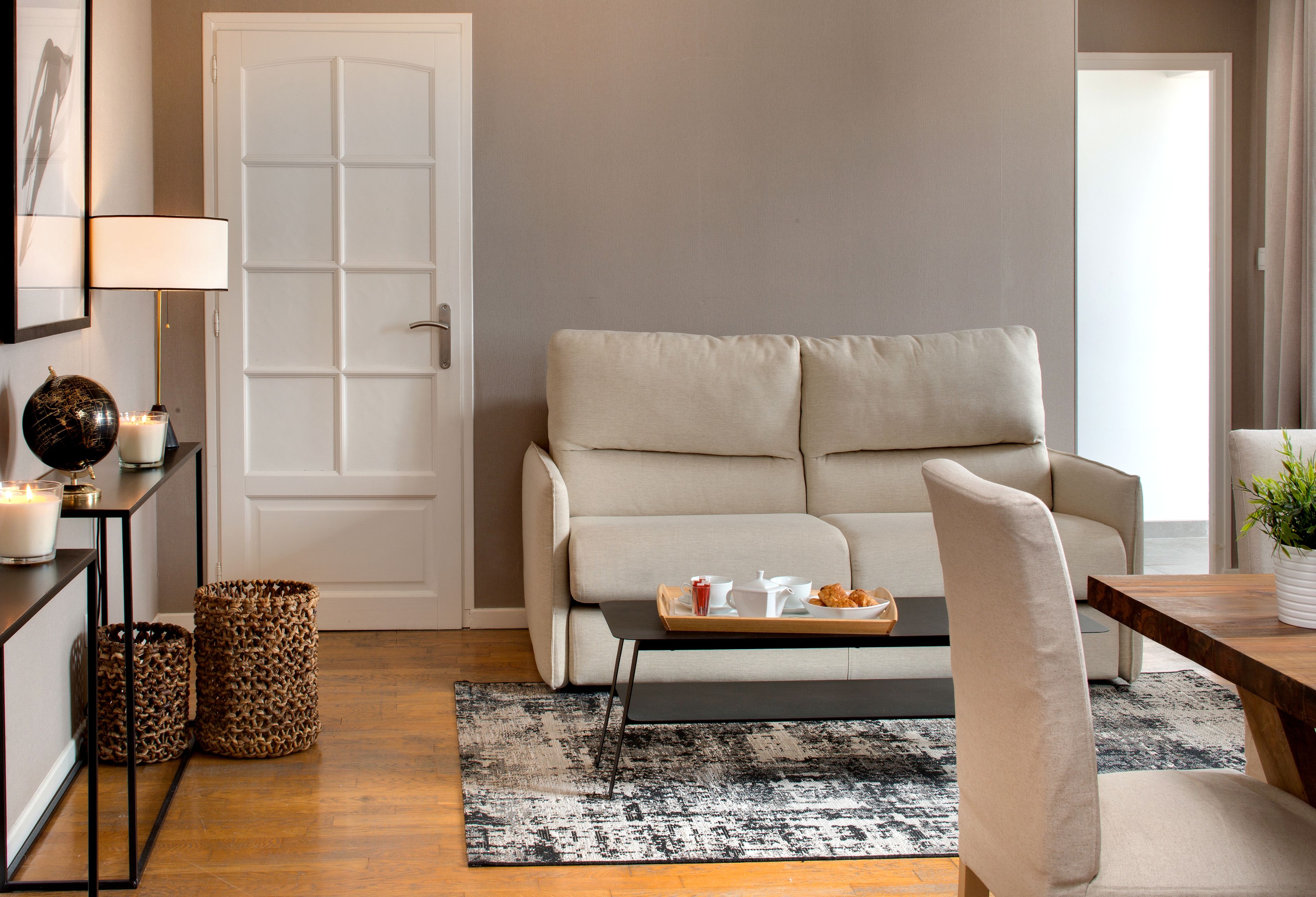 A living room with a beige sofa, a coffee table with a tray of items, a patterned rug, and a door in the background.