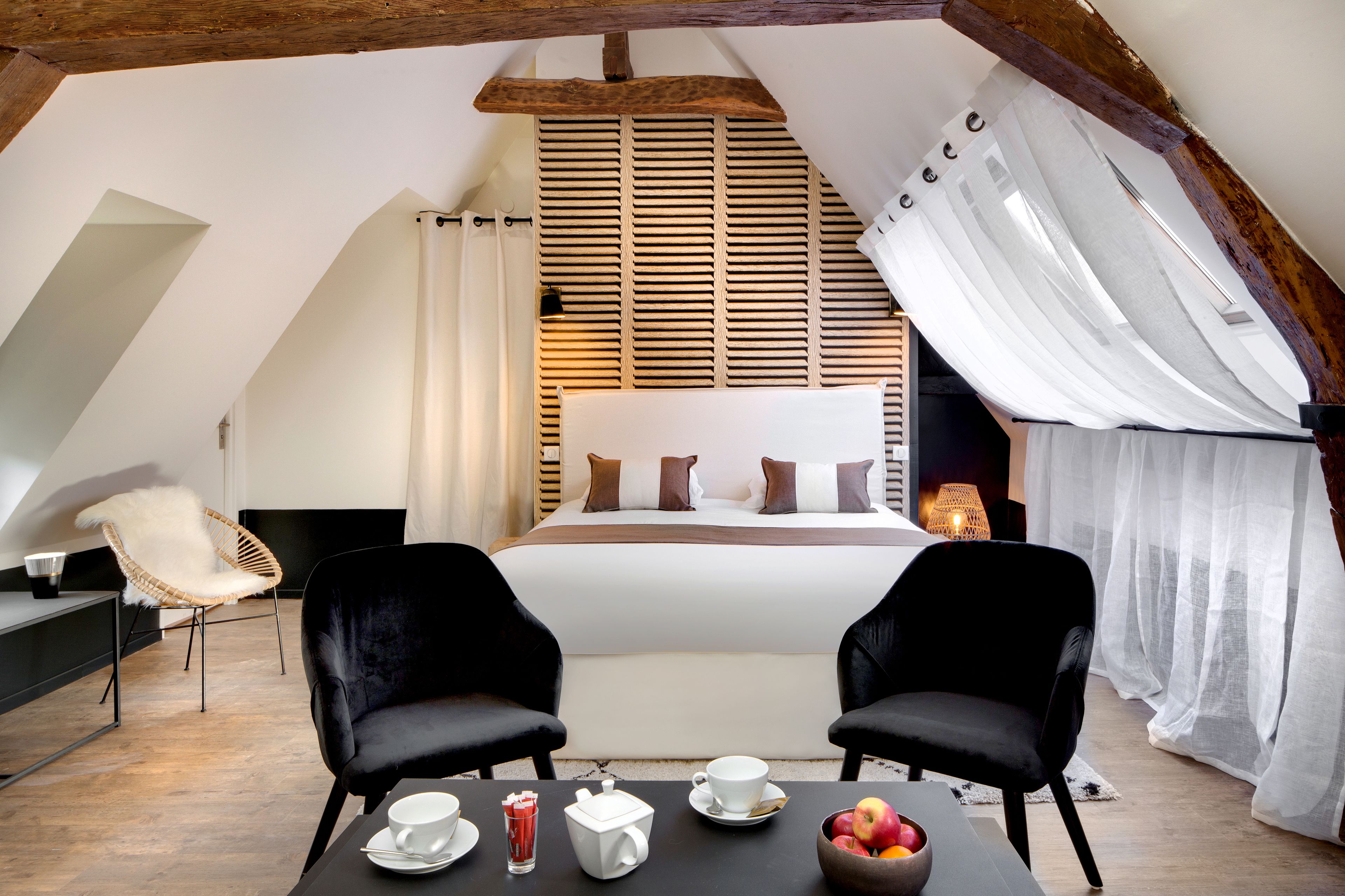 A cozy bedroom with a large bed, two black chairs, a table with breakfast items, and a wooden slatted window blind.