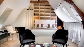 A cozy bedroom with a large bed, two black chairs, a table with breakfast items, and a wooden slatted window blind.
