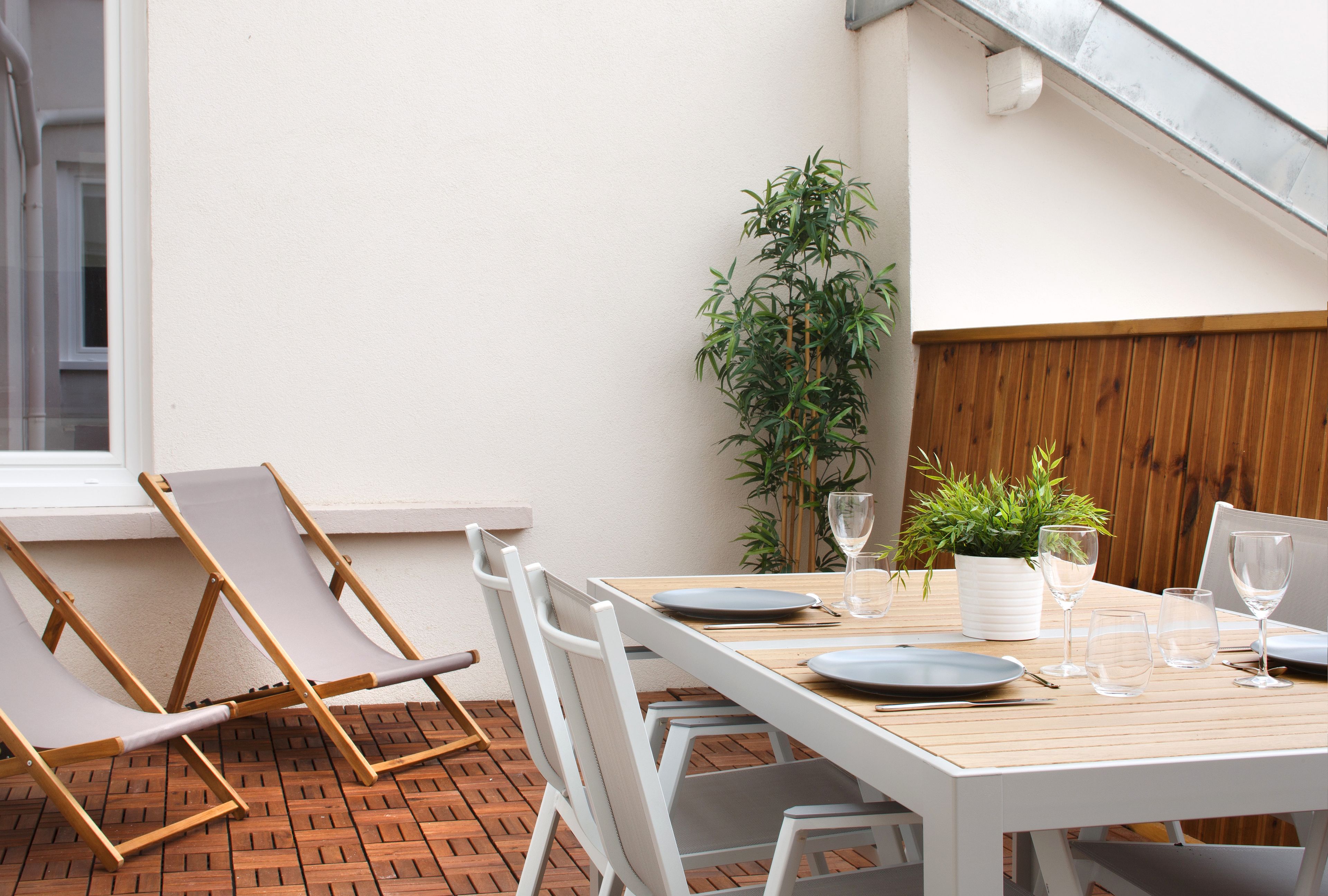 Un patio con una mesa de madera dispuesta para comer, sillas blancas y una planta en maceta.