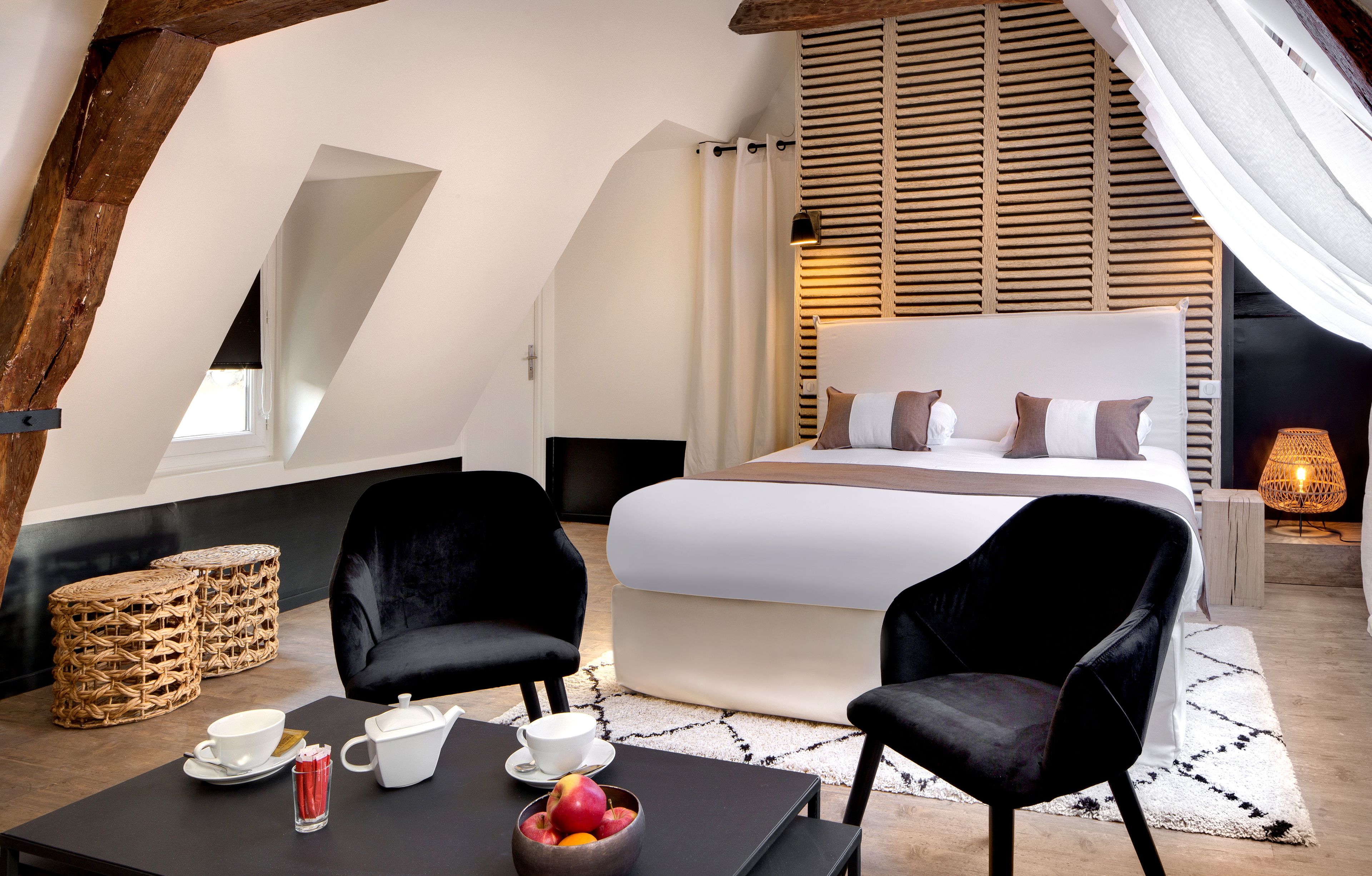 A bedroom with a large bed, black chairs, a dining table with a bowl of fruit, and a small table with cups and a teapot.