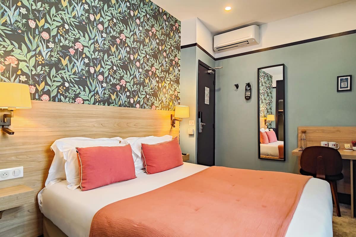 Ein Hotelzimmer mit einem großen Bett, zwei Nachttischlampen, einem hölzernen Kopfteil, floralem Wandpapier, einem Schreibtisch mit Stuhl und einem Spiegel.