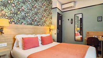 Quarto de hotel com uma cama grande, duas luminárias de cabeceira, uma cabeceira de madeira, papel de parede floral, uma escrivaninha com cadeira e um espelho.