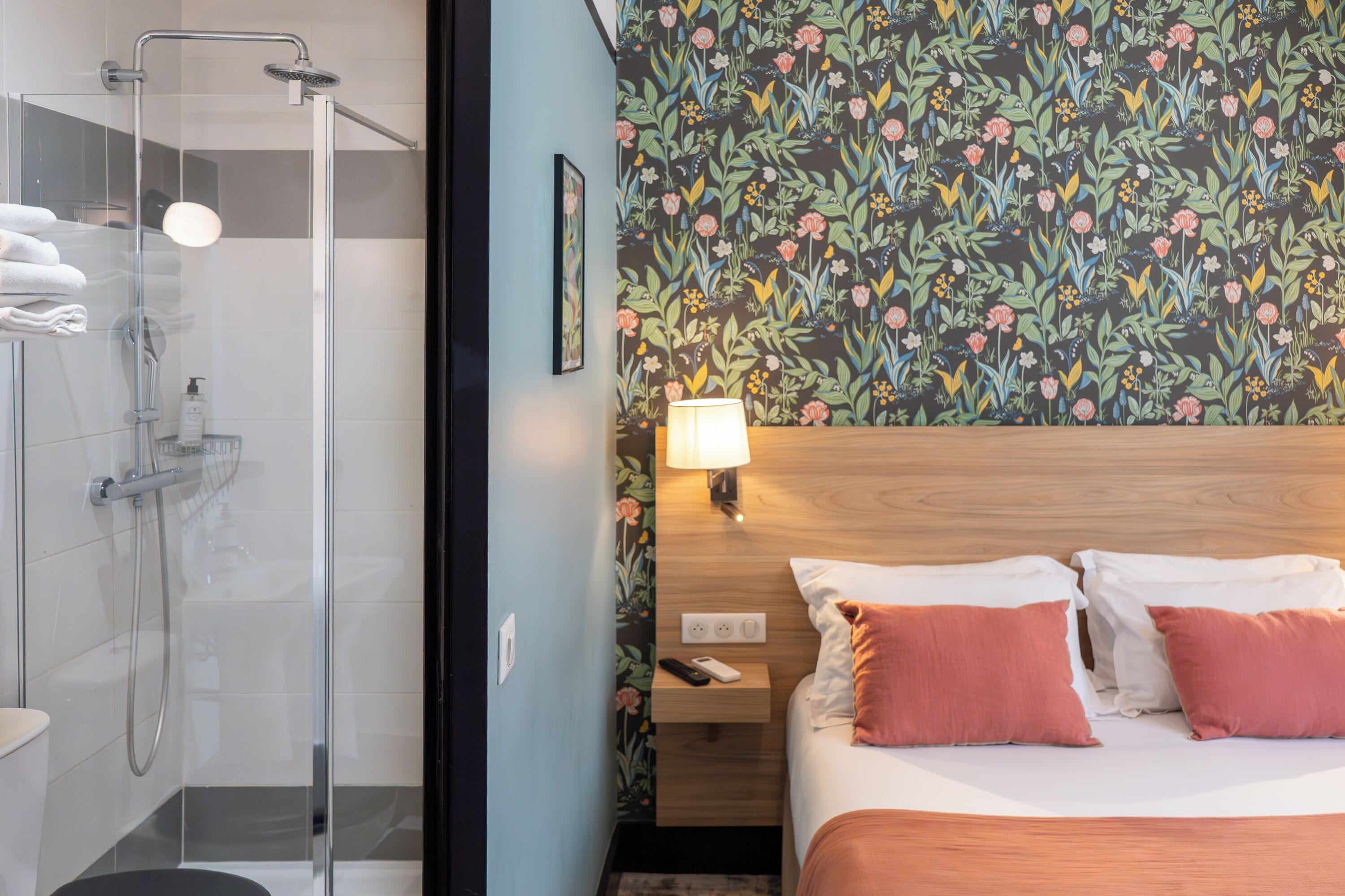 A hotel room with a shower, a bed with two pillows, a nightstand with a lamp, and a floral wallpaper.