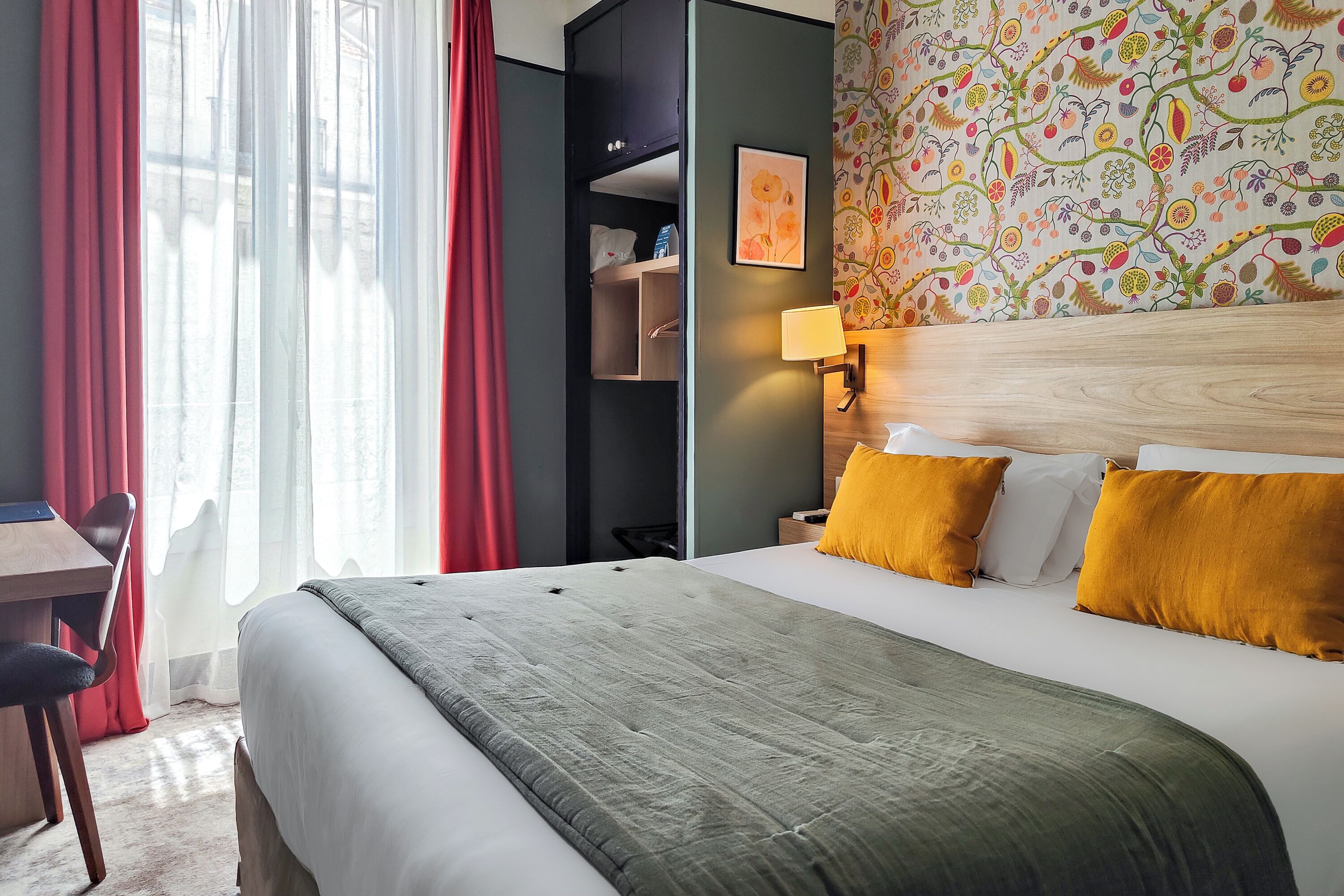 Camera d'albergo con un letto, una scrivania e una sedia. Si vede una finestra con tende, una parete con motivo floreale e una lampada a parete.