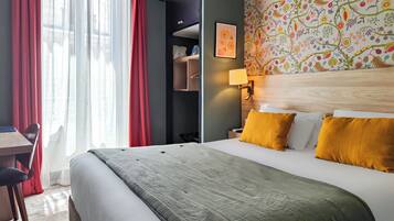 A hotel room with a bed, a desk, and a chair. There is a window with curtains, a wall with a floral pattern, and a lamp on the wall.