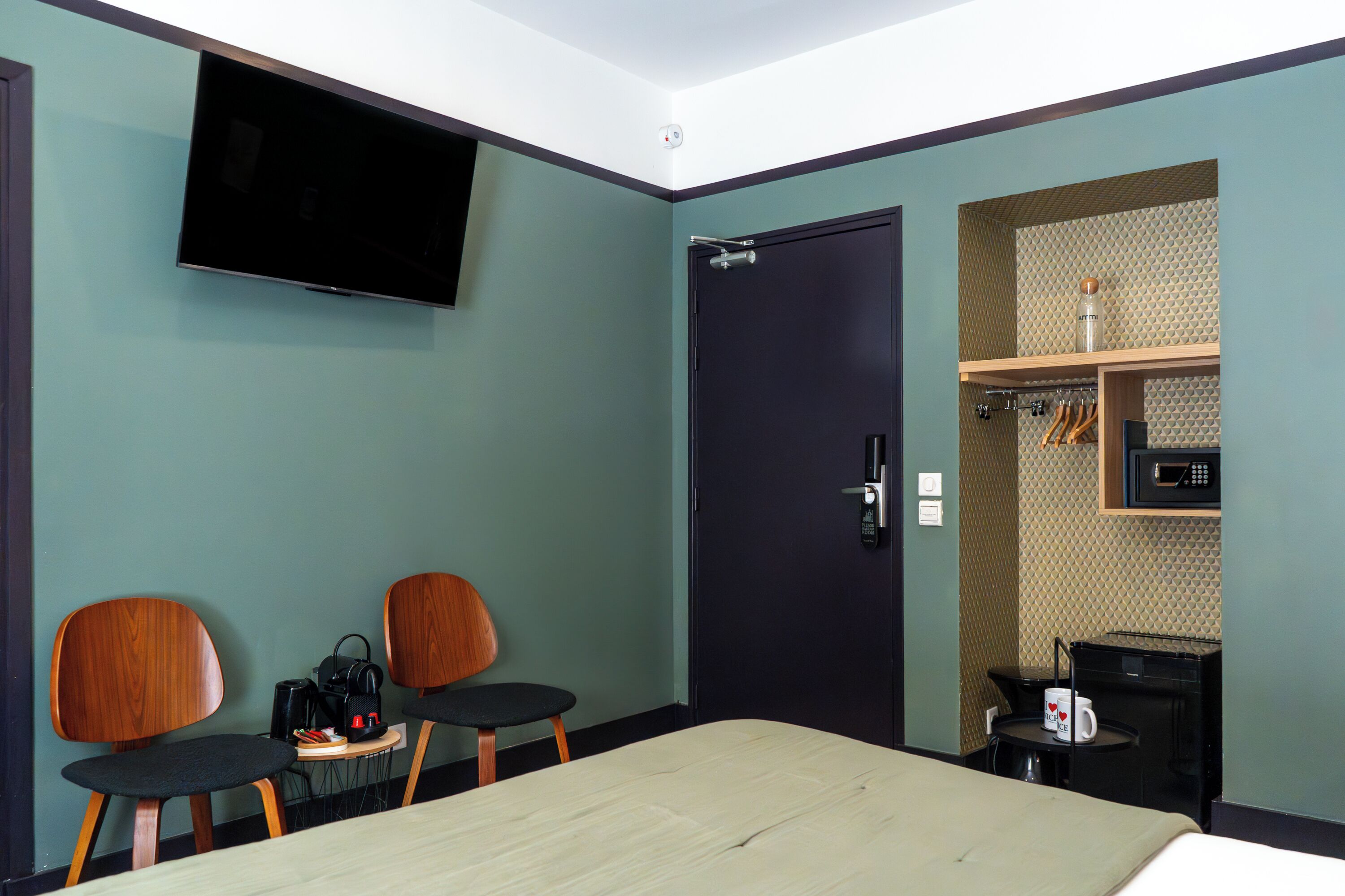 Habitación de hotel con una cama, dos sillas de madera, una mesita con tetera y cafetera, un televisor montado en la pared y una puerta con manija.