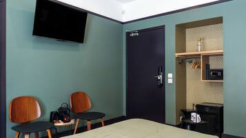 Habitación de hotel con una cama, dos sillas de madera, una mesita con tetera y cafetera, un televisor montado en la pared y una puerta con manija.