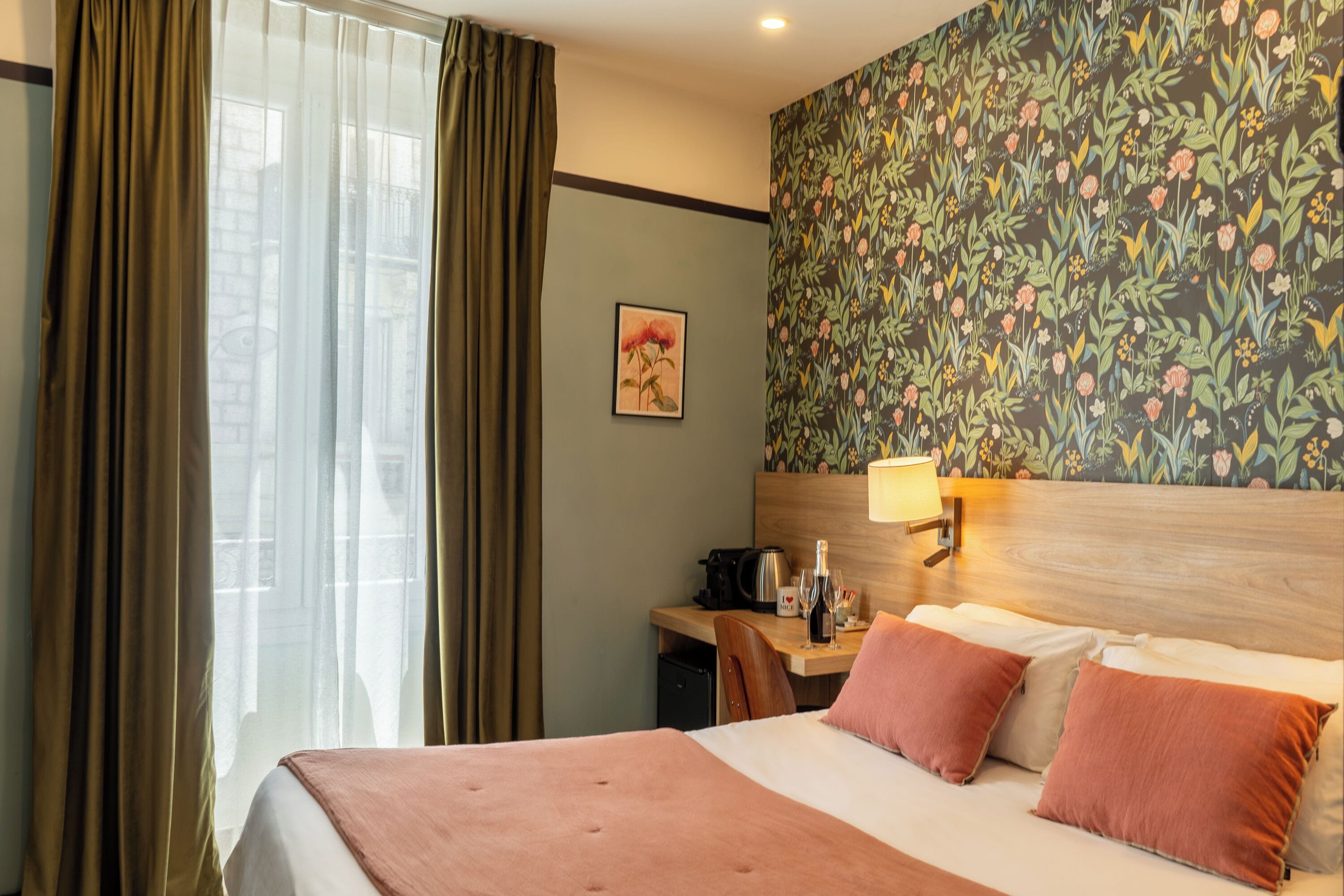 A hotel room with a bed, a desk with a lamp, a small refrigerator, and a floral wallpaper.