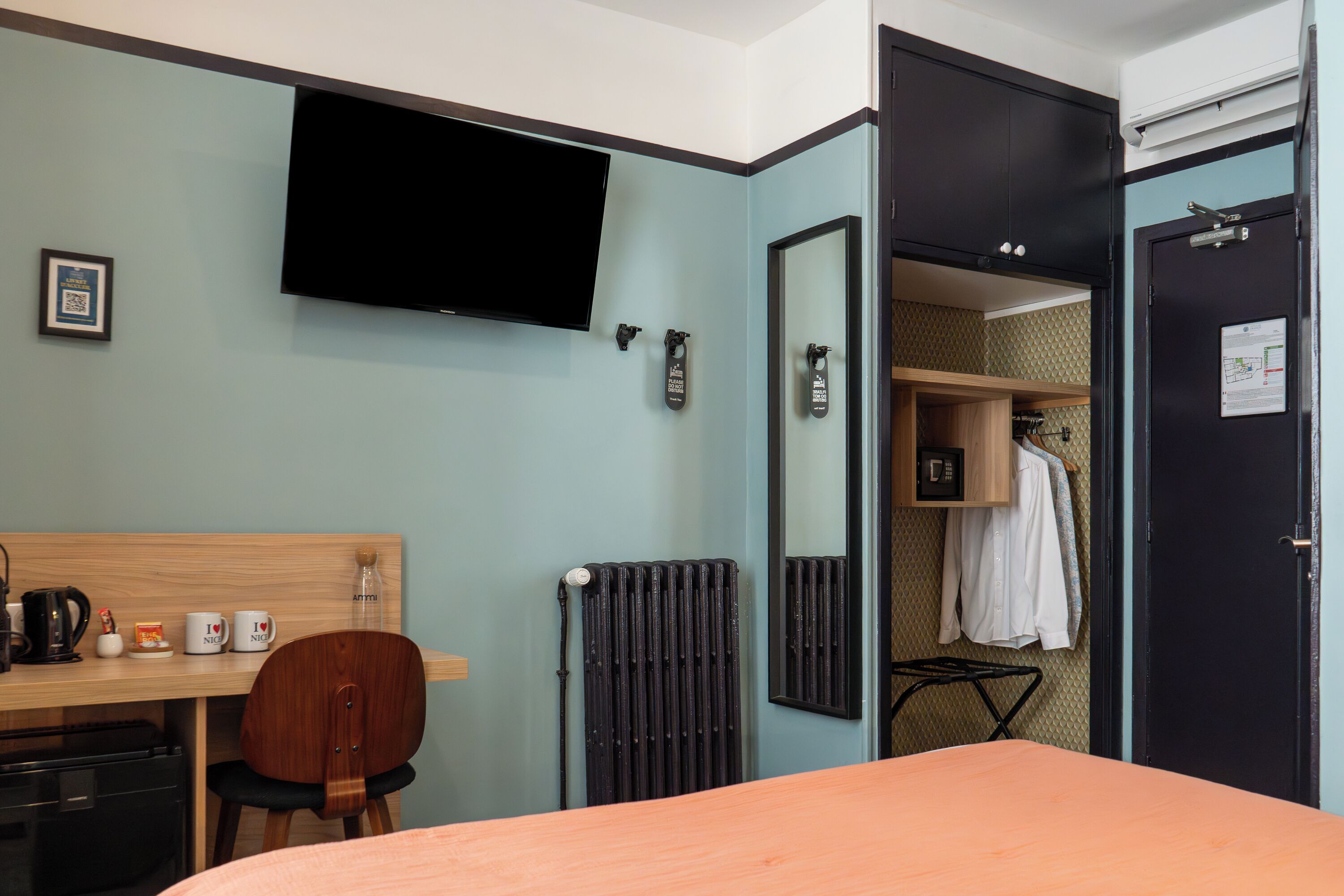 A hotel room with a bed, a desk with a chair, a television, a radiator, and a wardrobe.