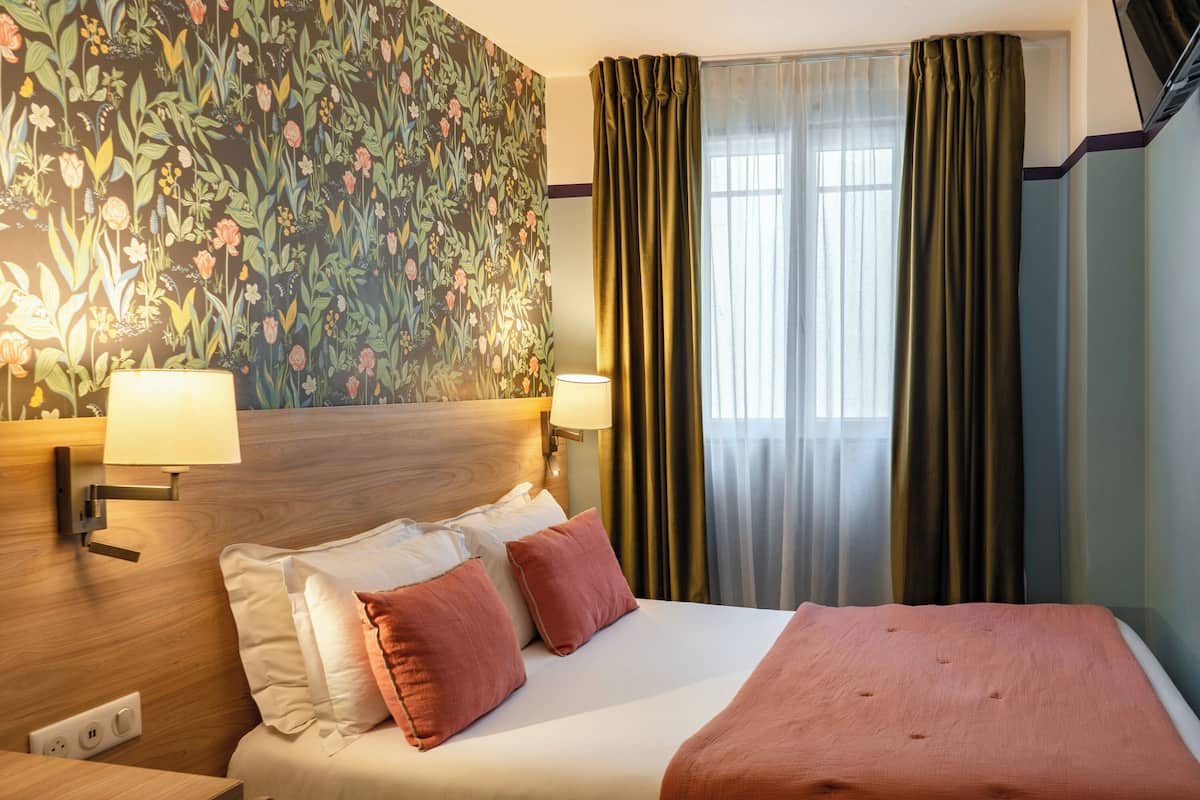 Ein Hotelzimmer mit einem großen Bett, zwei Wandlampen, blumigem Tapetenmuster und einem Fenster mit Vorhängen.