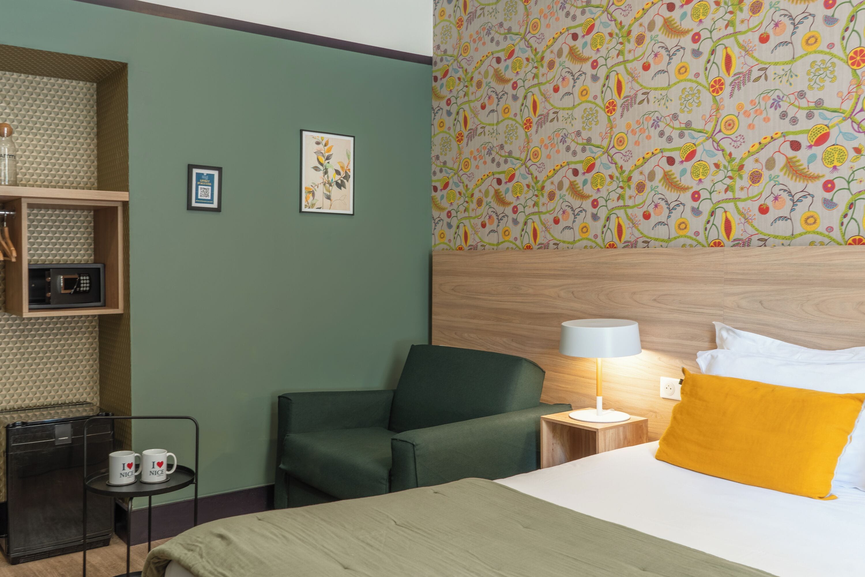 A hotel room with a green wall, a wooden headboard, a bed with white and yellow pillows, a small table with two mugs, a chair, and a patterned wallpaper.