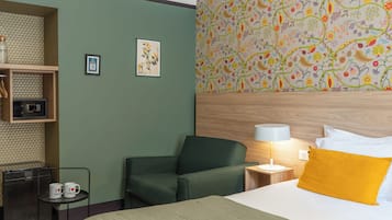 A hotel room with a green wall, a wooden headboard, a bed with white and yellow pillows, a small table with two mugs, a chair, and a patterned wallpaper.