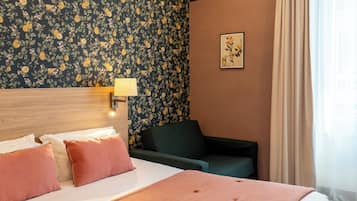 A hotel room with a bed, a nightstand, a lamp, a chair, a floral wallpaper, and a painting on the wall.