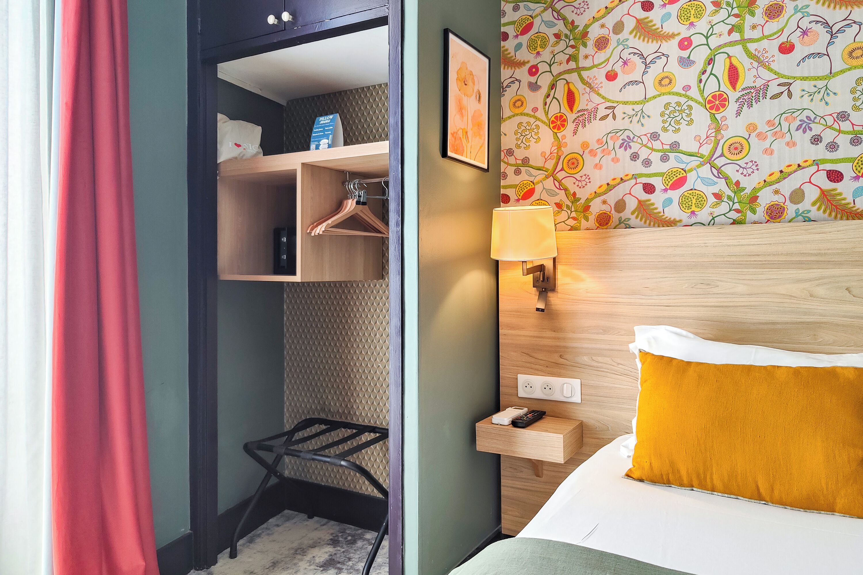 A hotel room with a bed, a nightstand, a wall-mounted lamp, a shelf with a phone and a remote, and a wardrobe with hanging clothes.