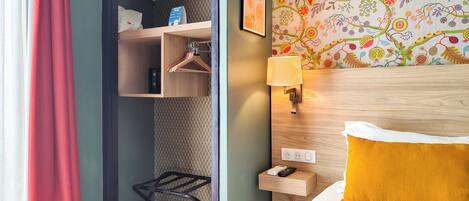 Habitación de hotel con cama, mesita de noche, lámpara de pared, estante con teléfono y control remoto, y armario con ropa colgada.