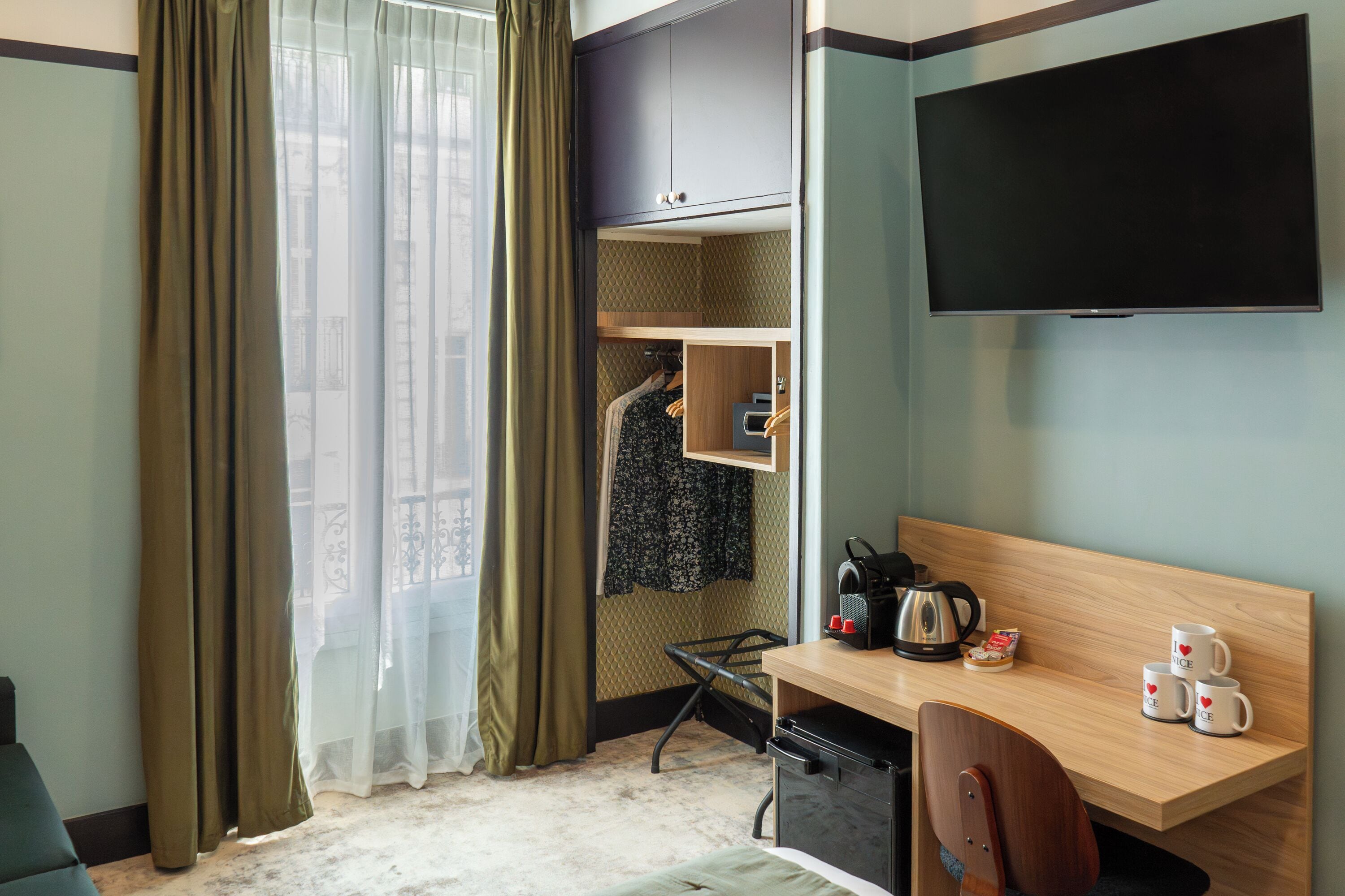 A hotel room with a bed, a desk with a chair, a TV, a wardrobe, and a window with curtains.