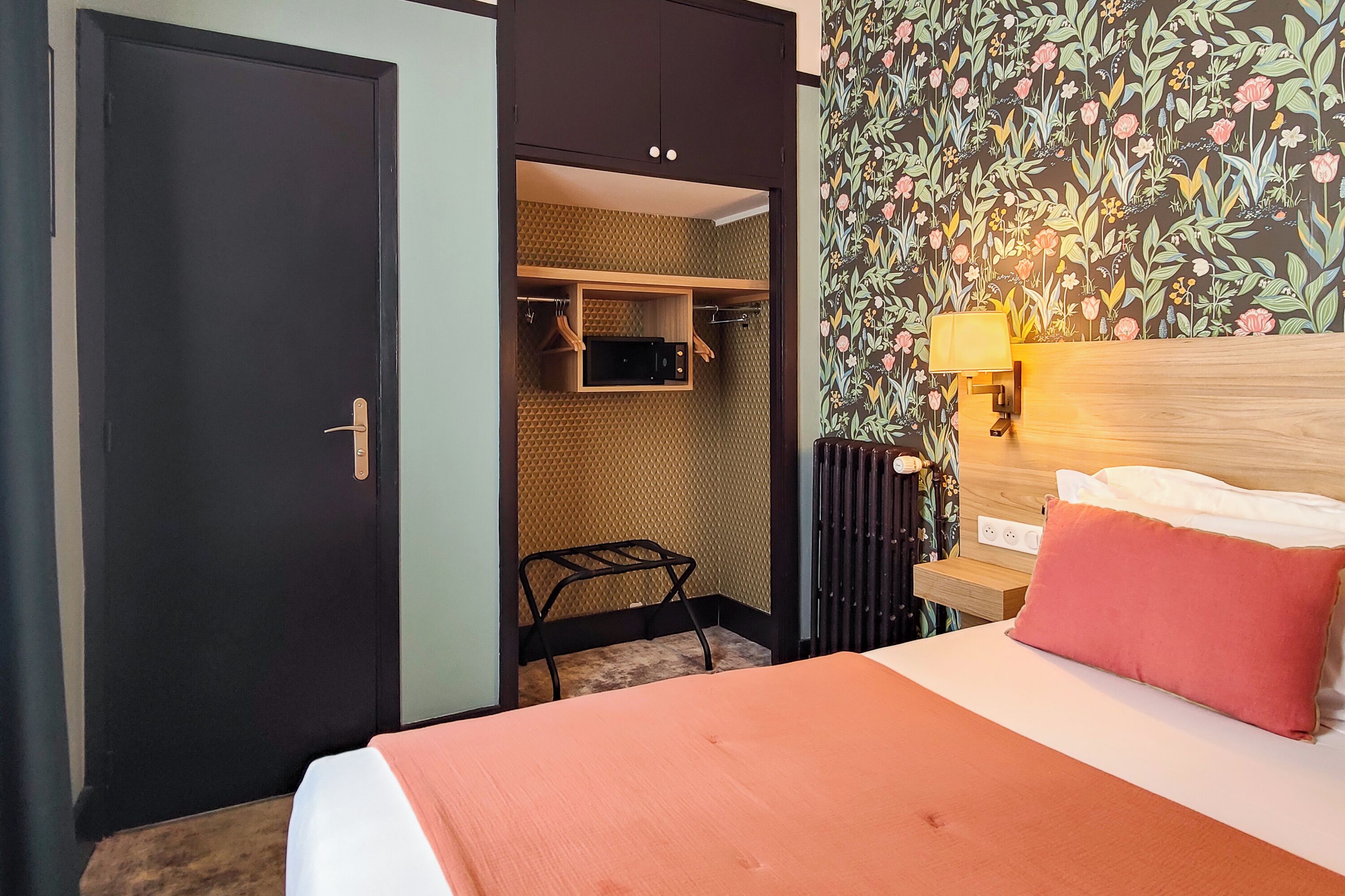 A hotel room with a bed, a nightstand with a lamp, a wardrobe, and a small seating area.