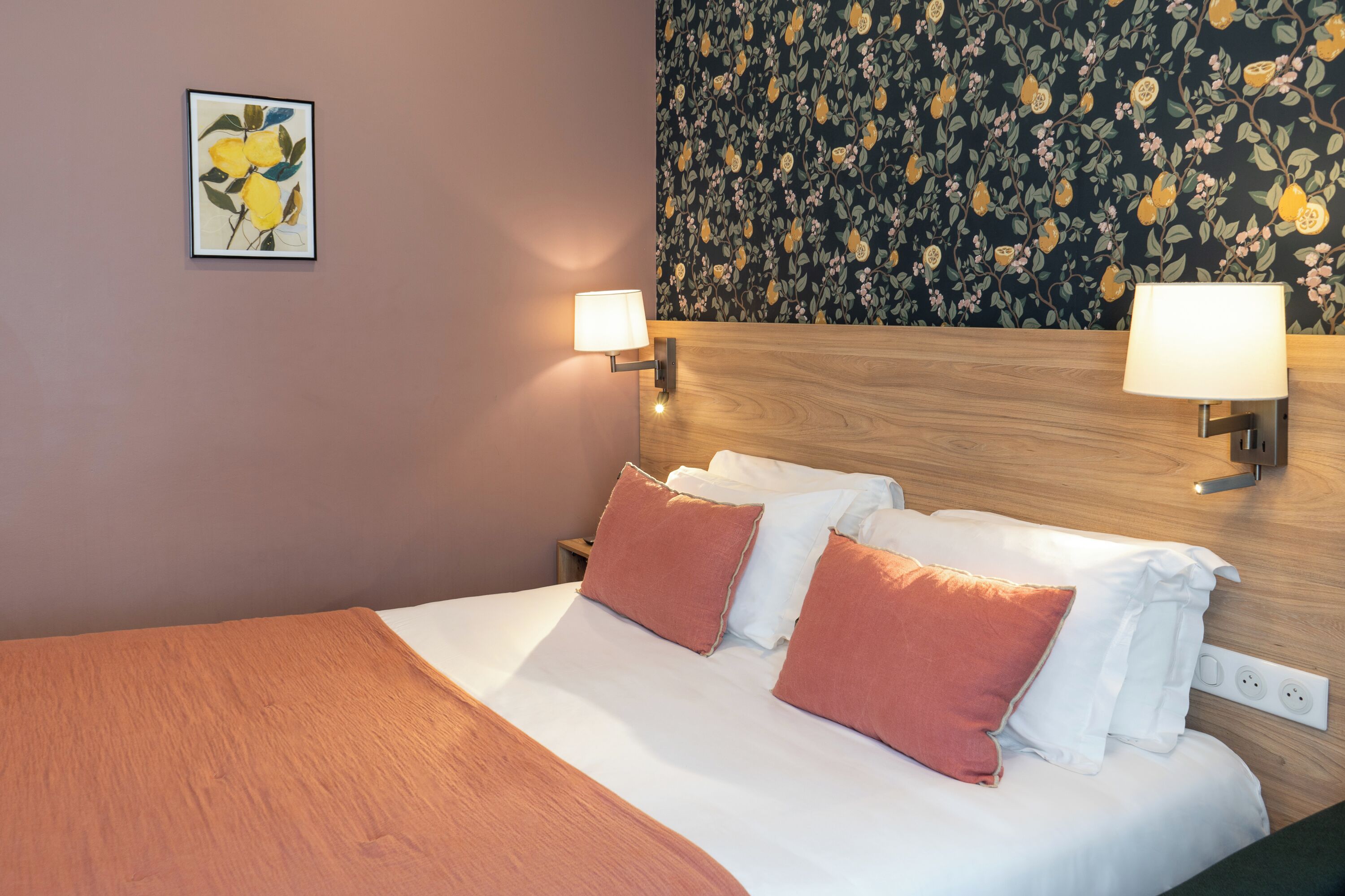 A neatly made bed with white and orange pillows, a wooden headboard, and two wall-mounted lamps.