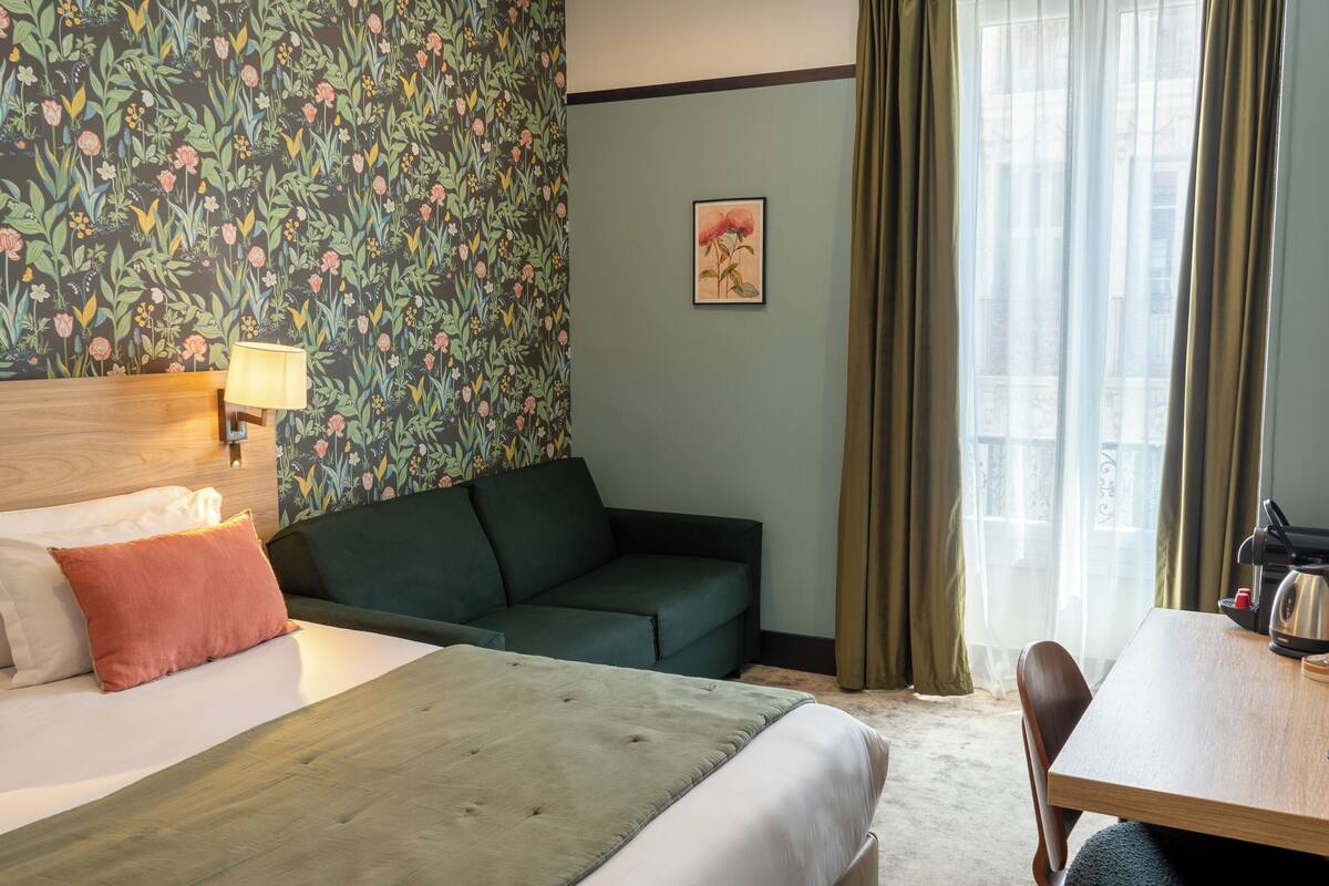 Ein Hotelzimmer mit einem Bett, einem grünen Sofa, einem Holztisch und einem Fenster mit Vorhängen.