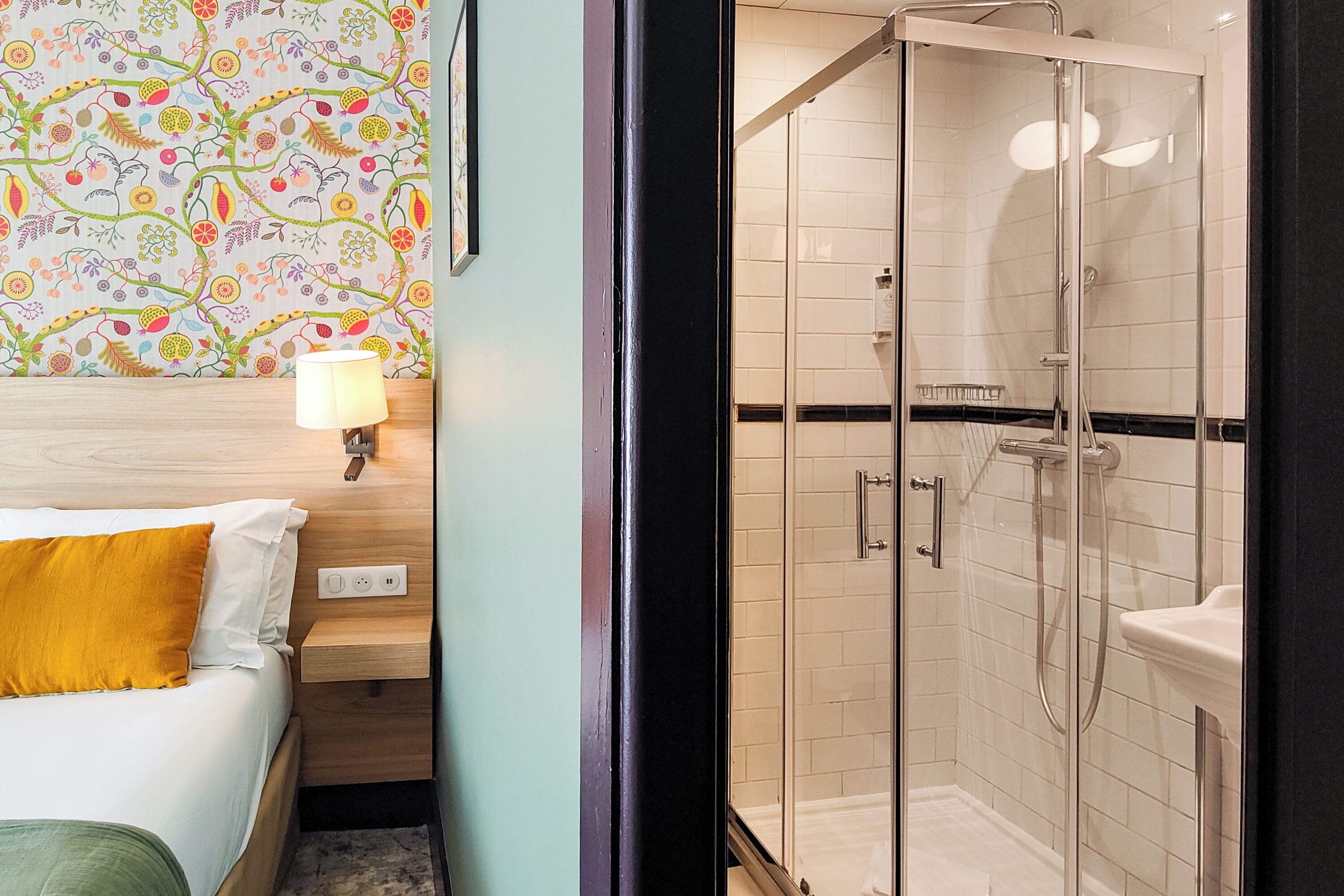 A hotel room with a bed, a nightstand, a lamp, a shower with glass doors, and a tiled wall.