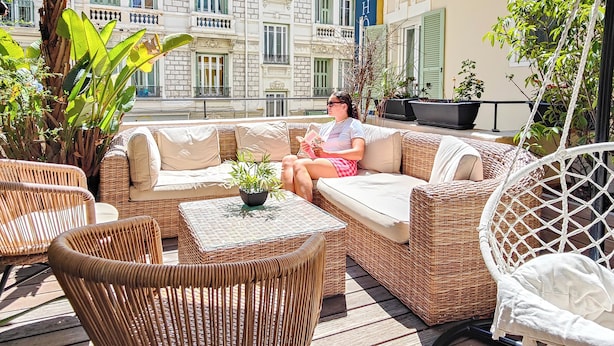 A wicker sofa with cushions, a glass-top table with a potted plant, and wicker chairs on an outdoor patio with a view of a building with balconies and shutters. - Hôtel de France, un hôtel AMMI (Nice)