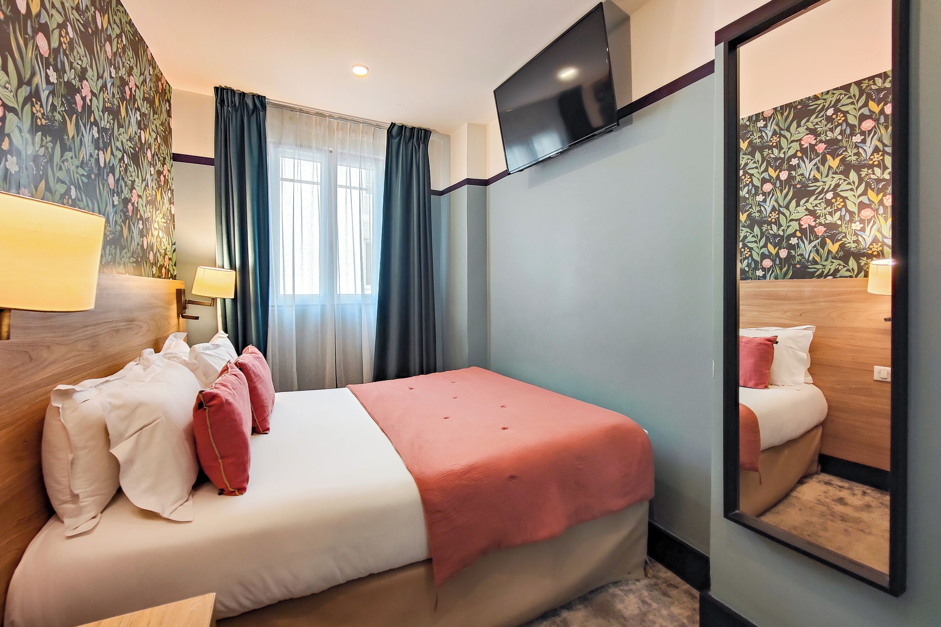 A hotel room with a bed, a TV mounted on the wall, a mirror, and a window with curtains.