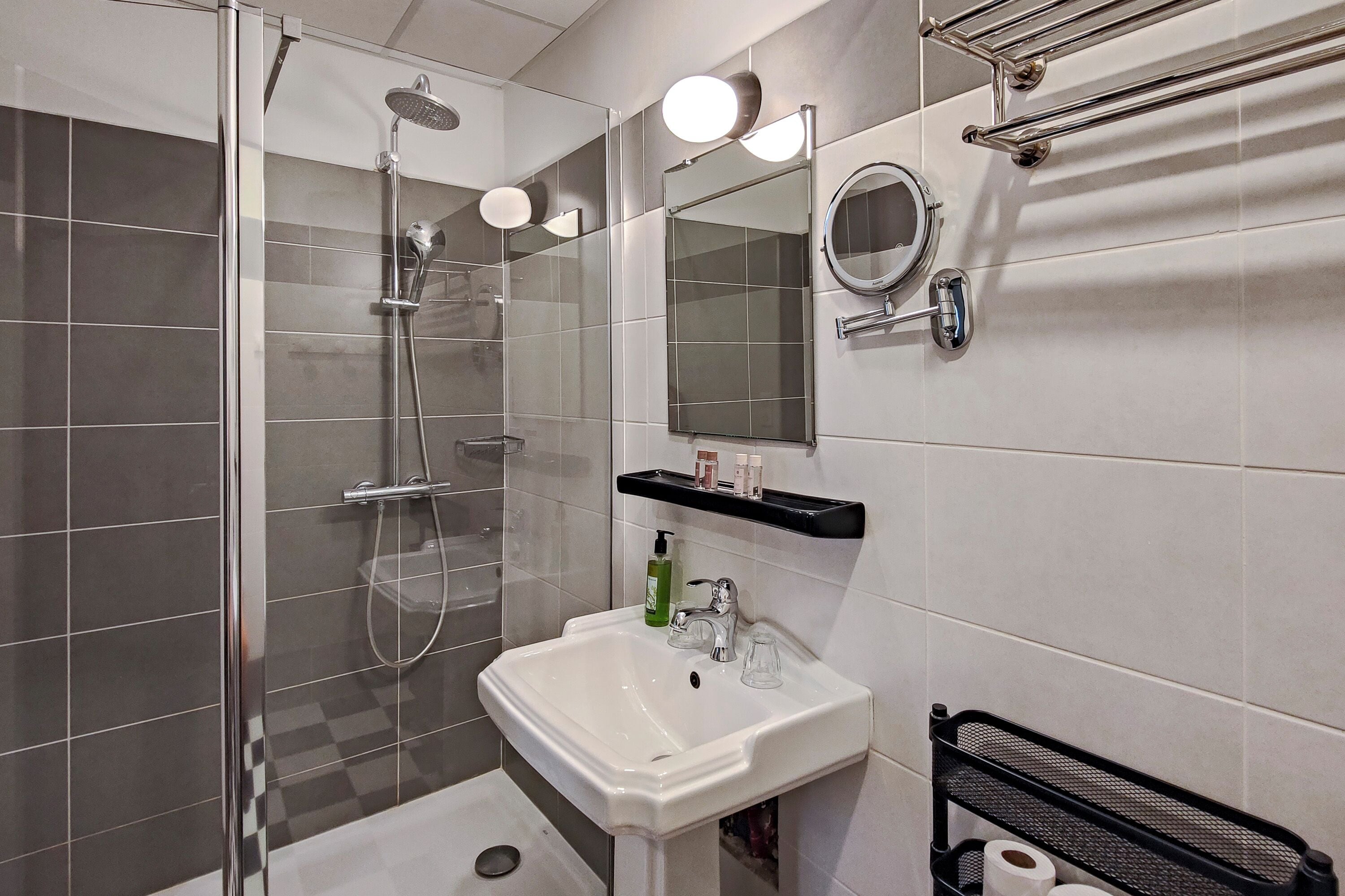 Un baño moderno con ducha de vidrio, un lavamanos blanco, un espejo y un toallero negro.