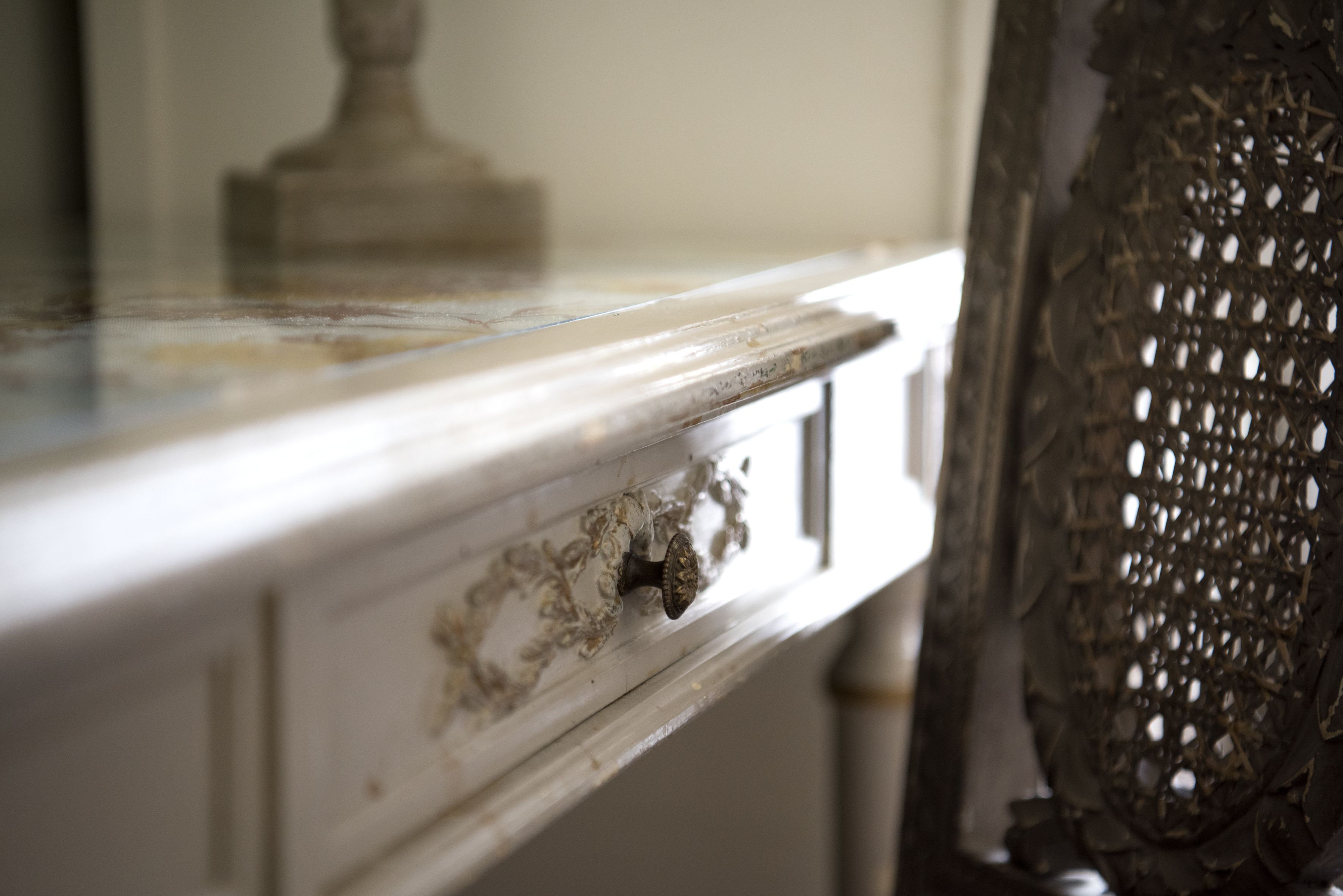 A close-up of a white dresser with a drawer handle, a decorative metal piece, and a lace curtain.