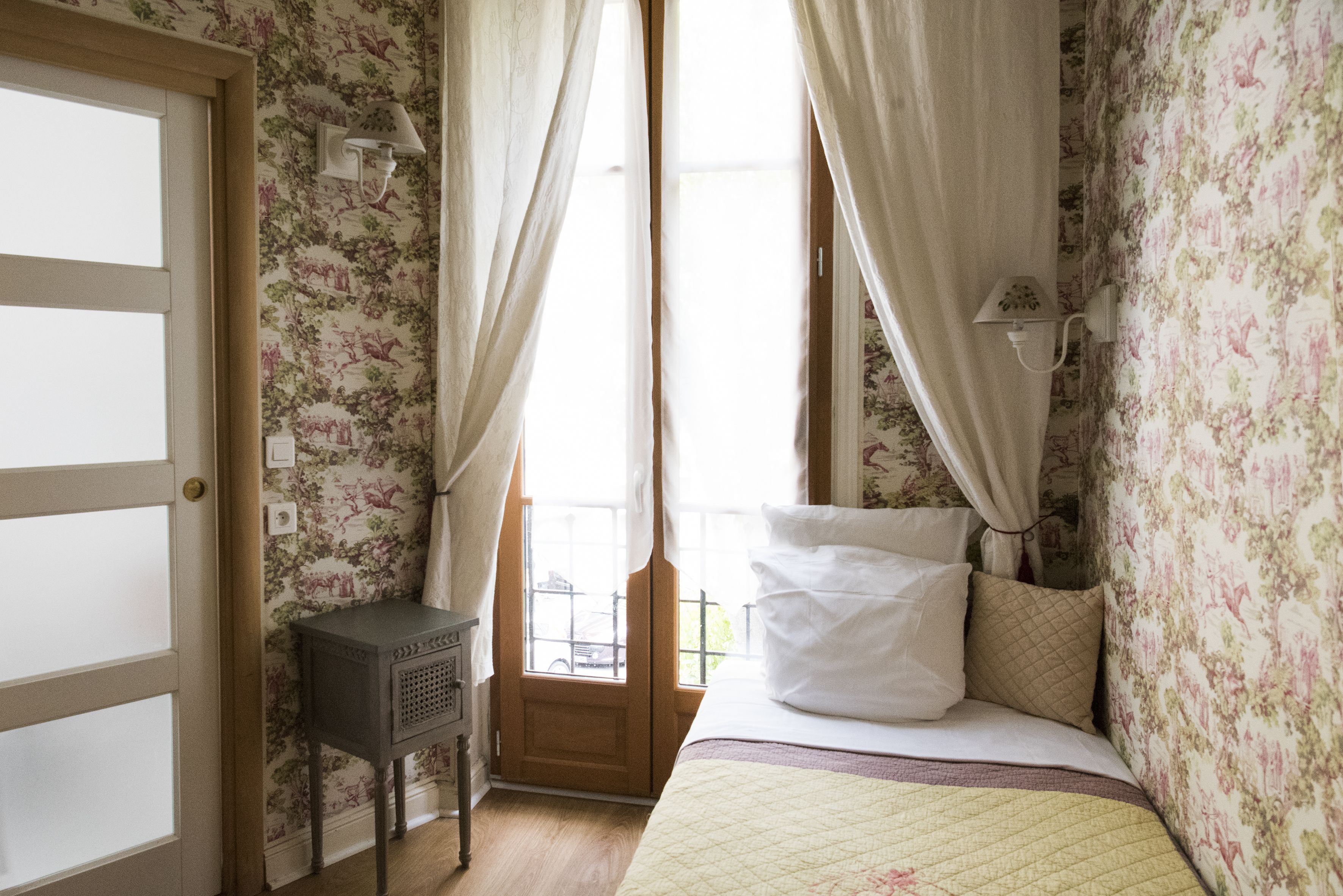 Een slaapkamer met bloemenbehang, een bed met witte en gemêleerde kussens, een nachtkastje en een raam met gordijnen.