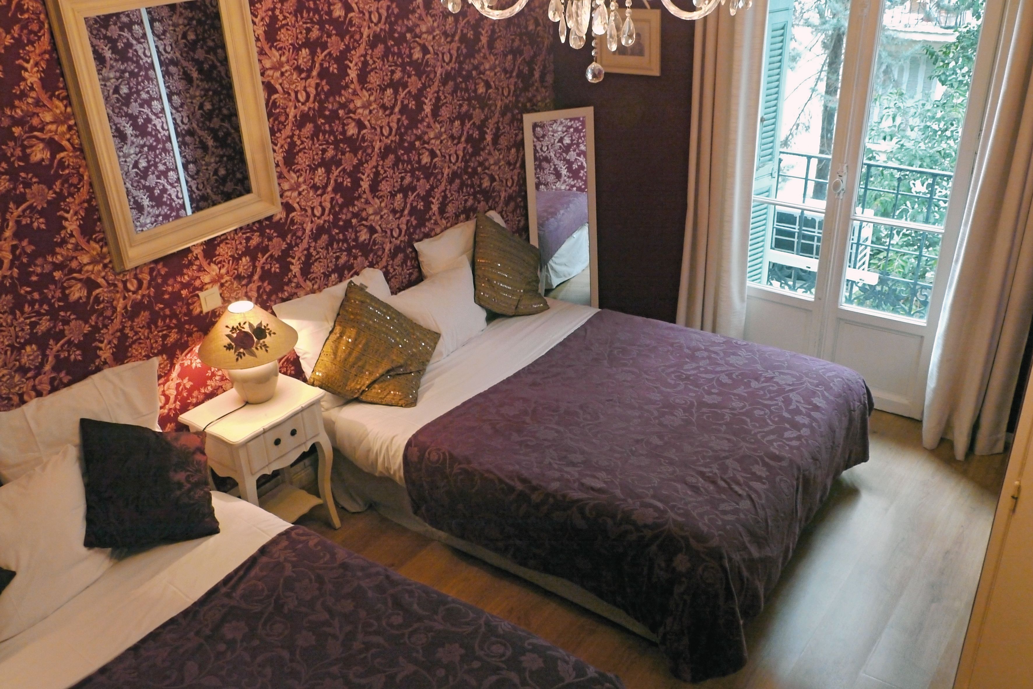 A bedroom with two beds, a nightstand with a lamp, and a window with a view of greenery.
