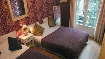 A bedroom with two beds, a nightstand with a lamp, and a window with a view of greenery.