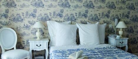 A bedroom with a bed, white nightstands, a white chair, and a blue patterned bedspread.