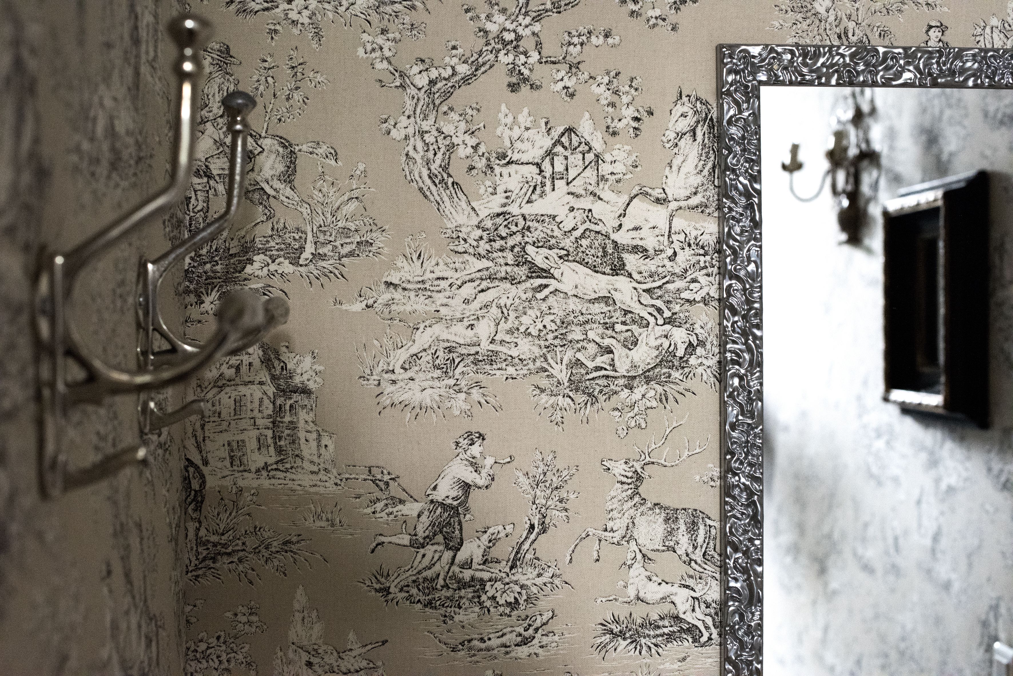 Una habitación con un papel tapiz detallado que muestra una escena campestre, un espejo con marco decorativo y una lámpara fijada a la pared.