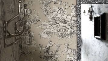 Una habitación con un papel tapiz detallado que muestra una escena campestre, un espejo con marco decorativo y una lámpara fijada a la pared.