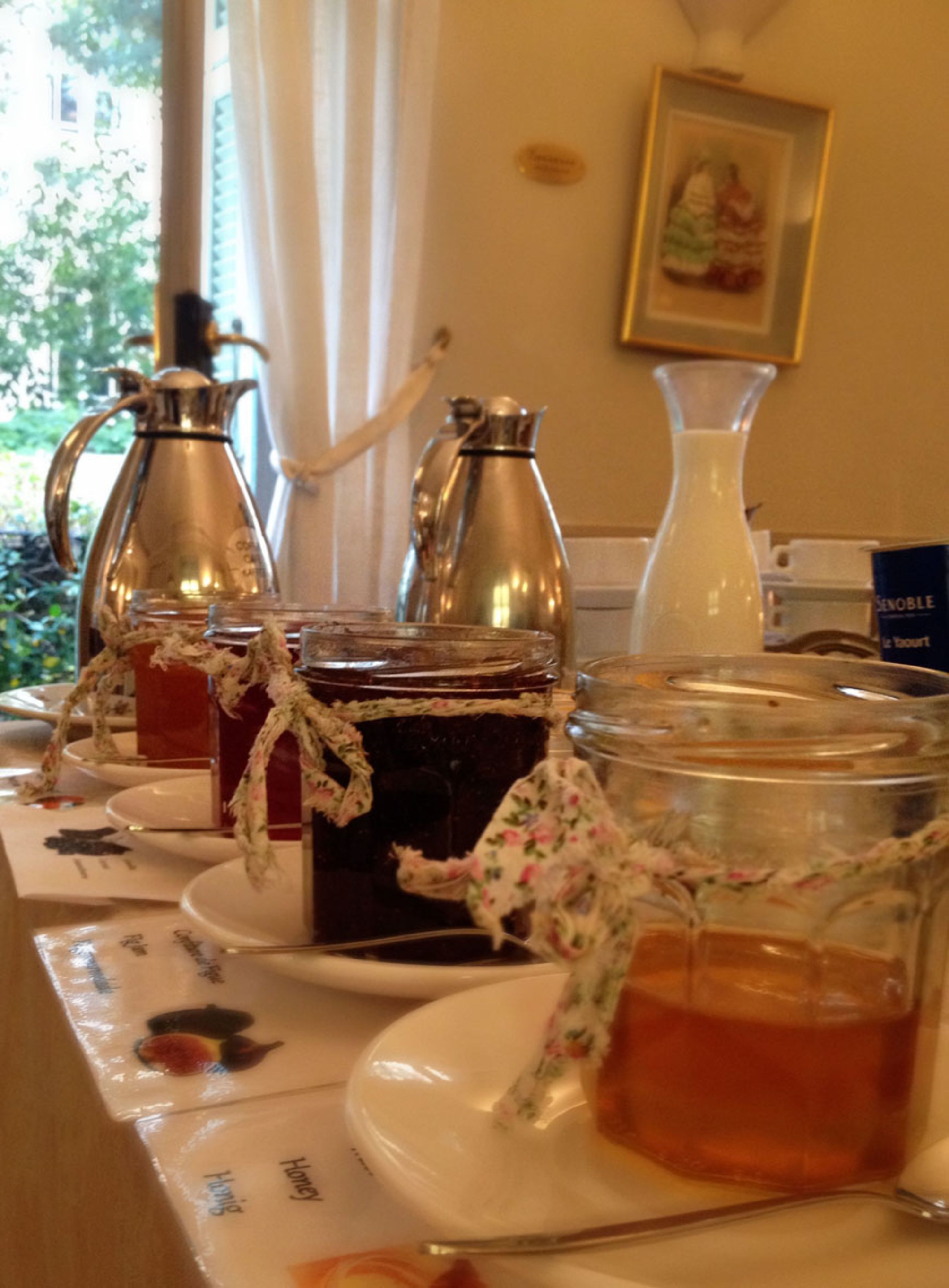 A table with various containers of honey and tea, including a glass jar and metal pitchers, set on a table with a floral tablecloth.