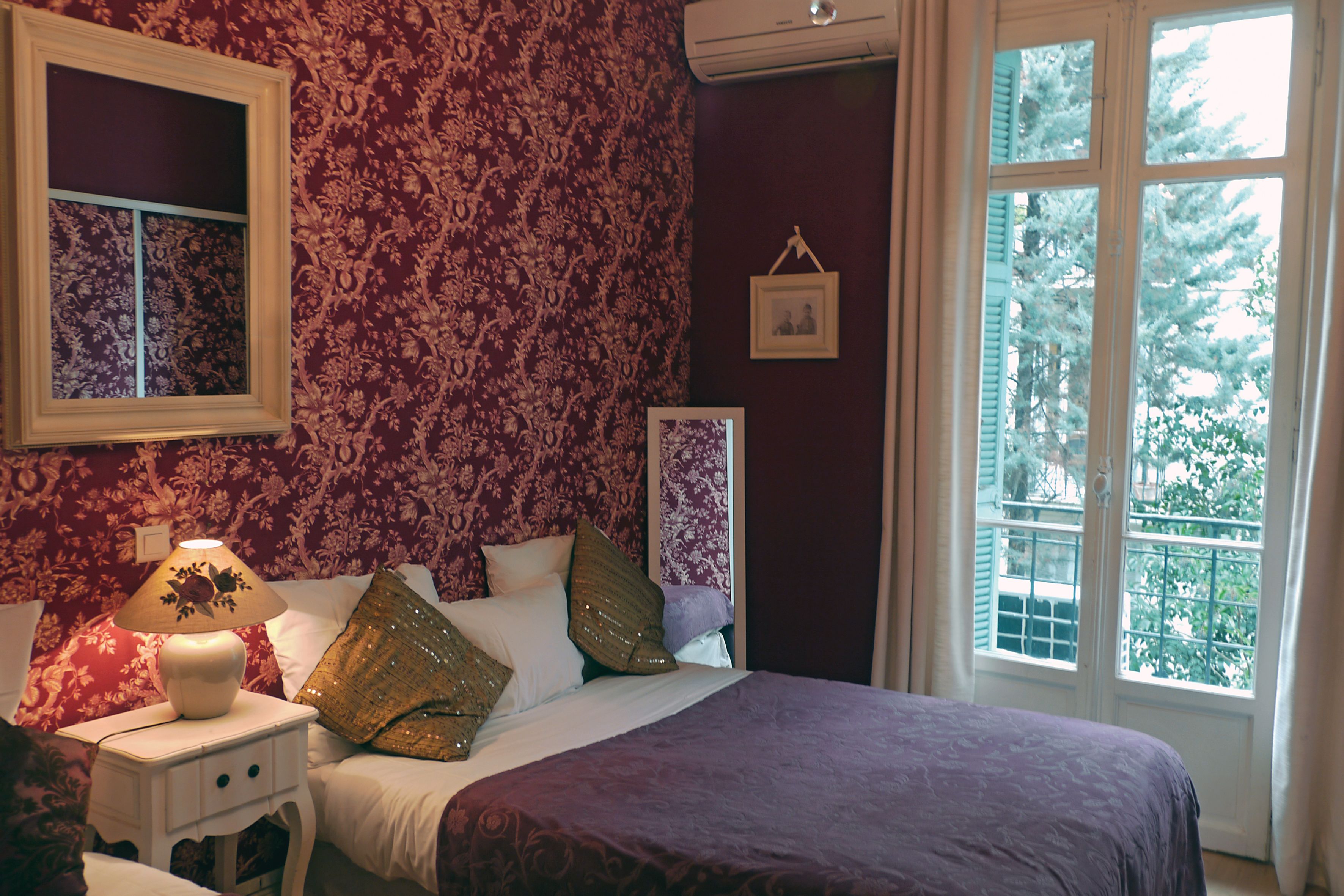 Ein Schlafzimmer mit einem großen Bett, einem Nachttisch mit Lampe, einem Fenster mit Blick auf Bäume und einer Wand mit floralem Wandpapier.