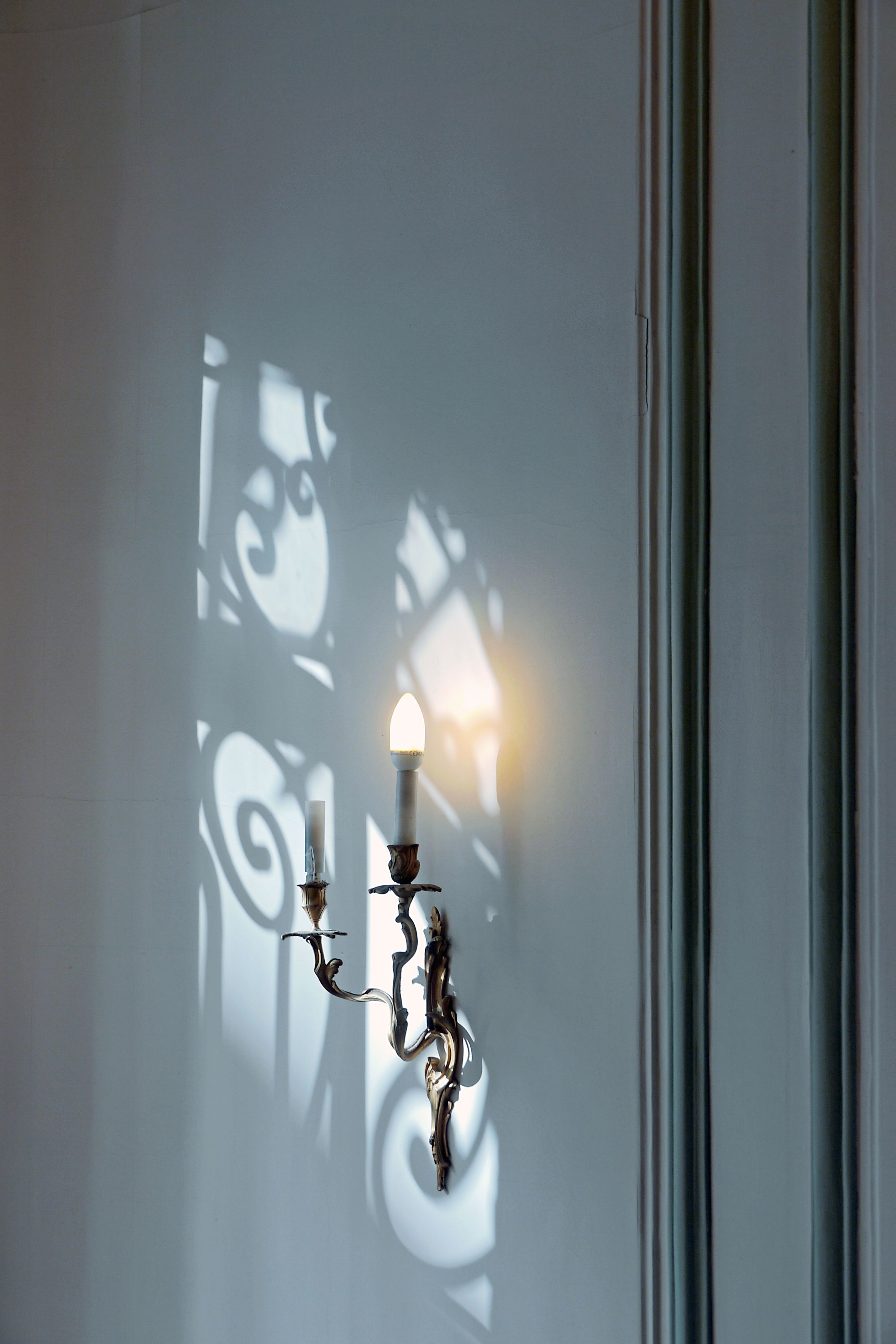 A wall-mounted light fixture with two candles, casting shadows of ornate designs on a plain wall.